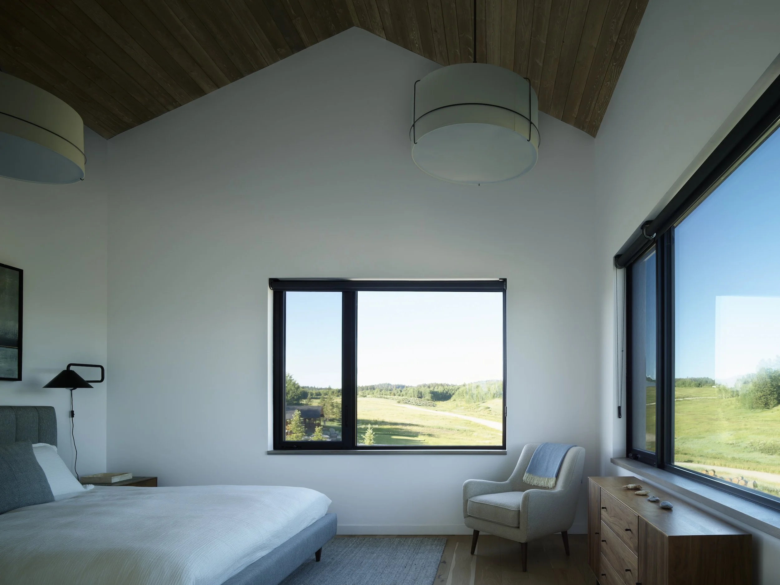 A modern bedroom with white walls, wooden ceiling, a large bed with white bedding, a white armchair with a blanket, and windows showing a green landscape.