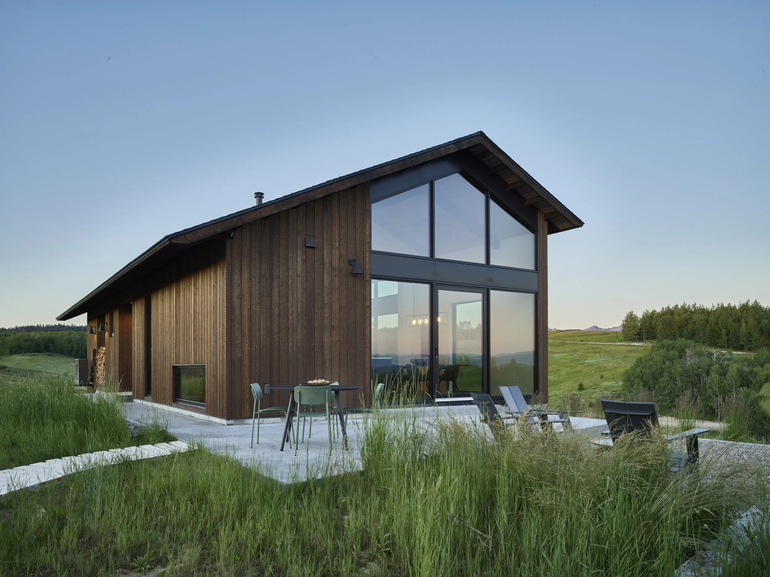 Modern house with large glass windows and wooden exterior, situated in a grassy landscape with outdoor furniture.