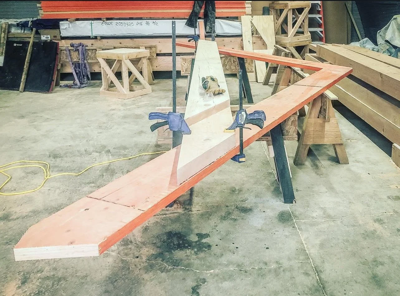 Woodworking workshop with a long wooden piece clamped to a sawhorse, featuring a geometric pattern painted on it, surrounded by various tools and materials.