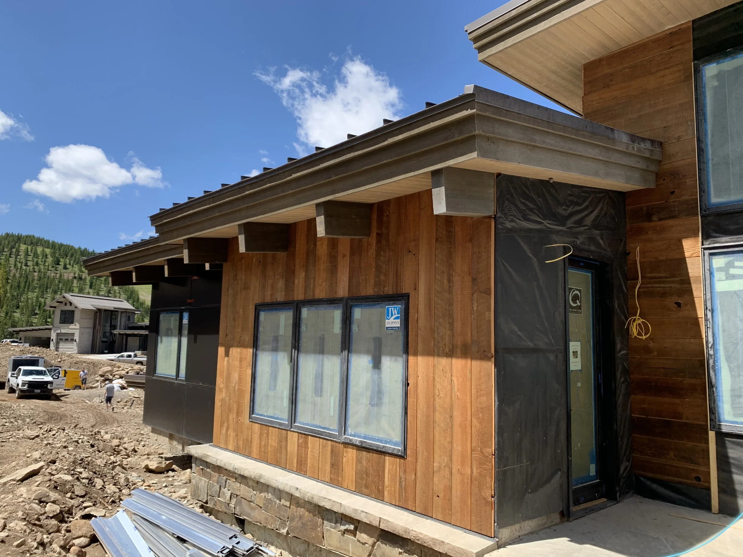 Construction site of a modern house with wood siding, large windows, and a stone foundation, under a blue sky with white clouds.