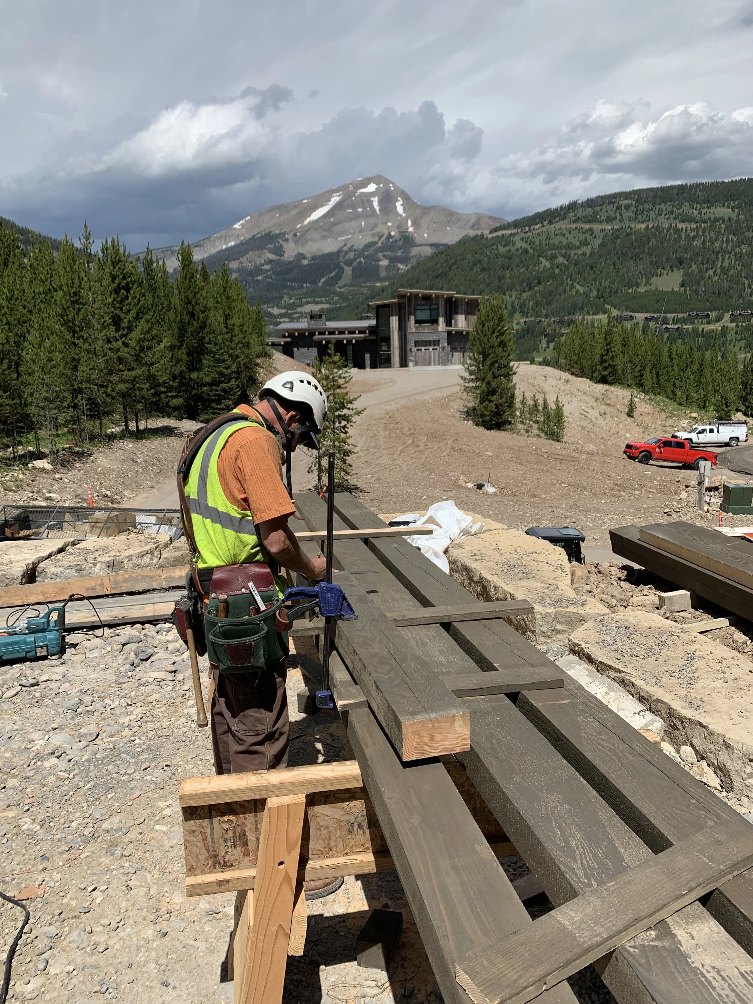 A construction worker wearing a yellow safety vest and a white helmet is working on a wooden structure outdoors with a mountain and cloudy sky in the background.