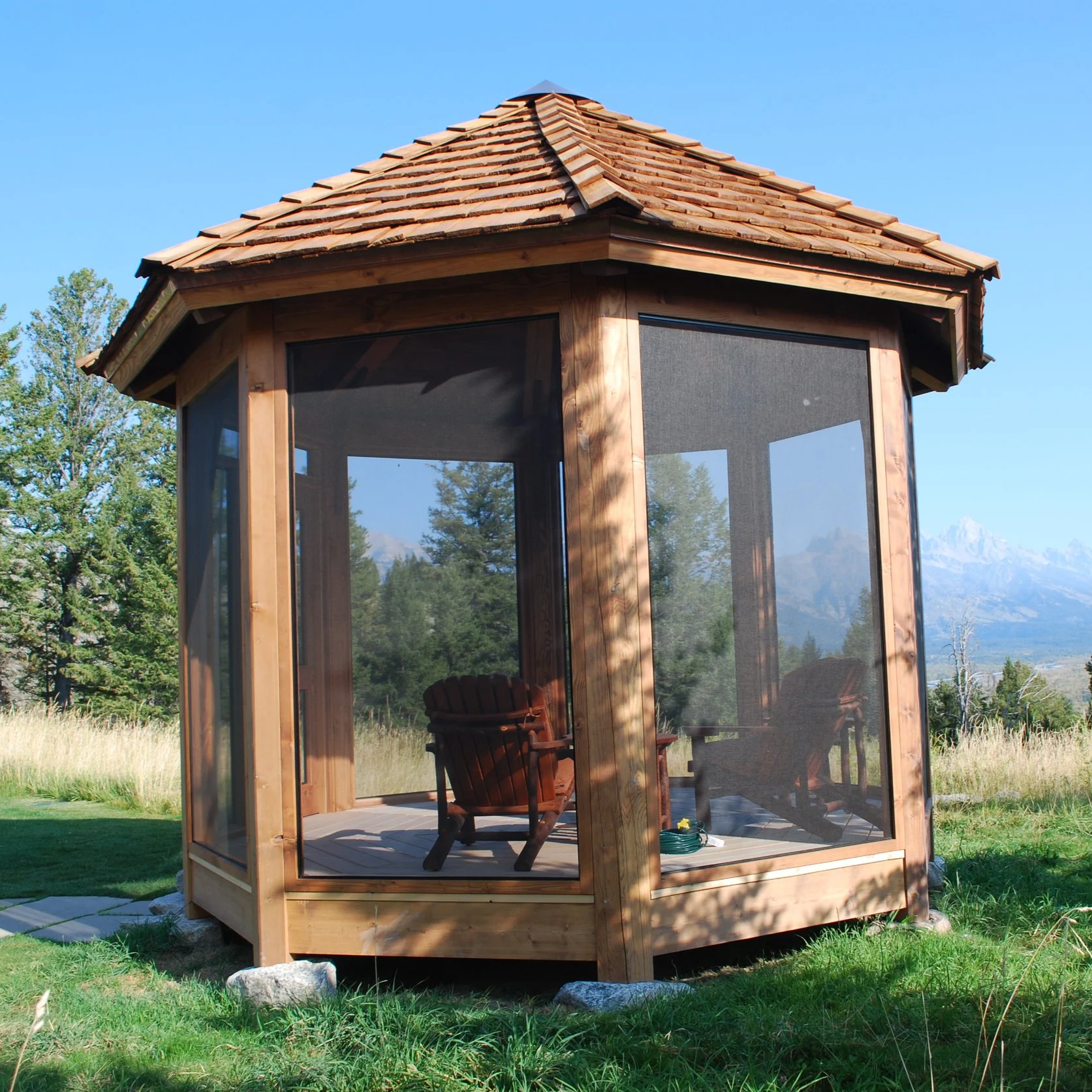 Wooden gazebo with screened sides, Adirondack chairs inside, set in a grassy area with trees and mountains in the background.