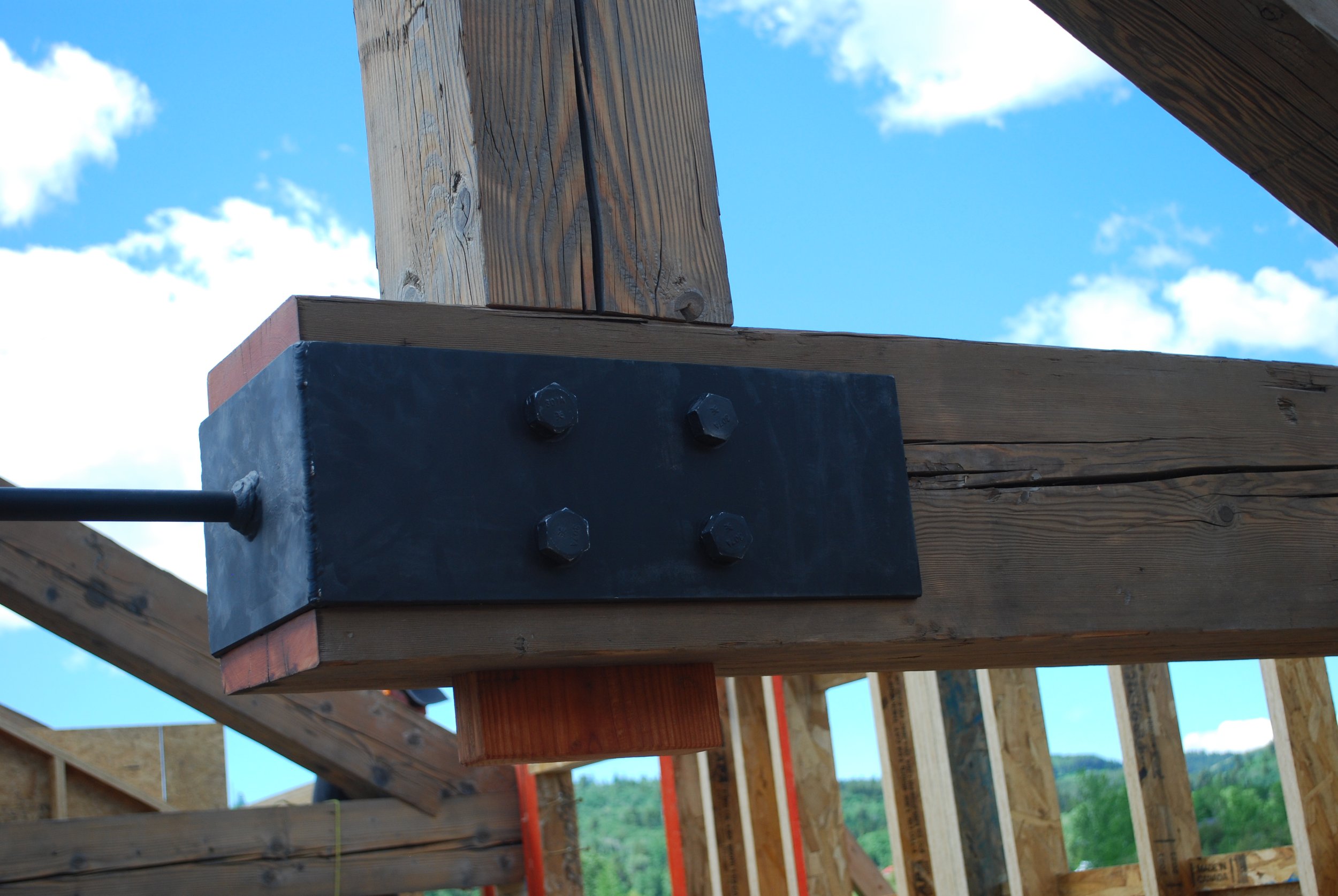 Close-up of a steel beam connector attached to wooden beams in a construction site, with blue sky and trees in the background.