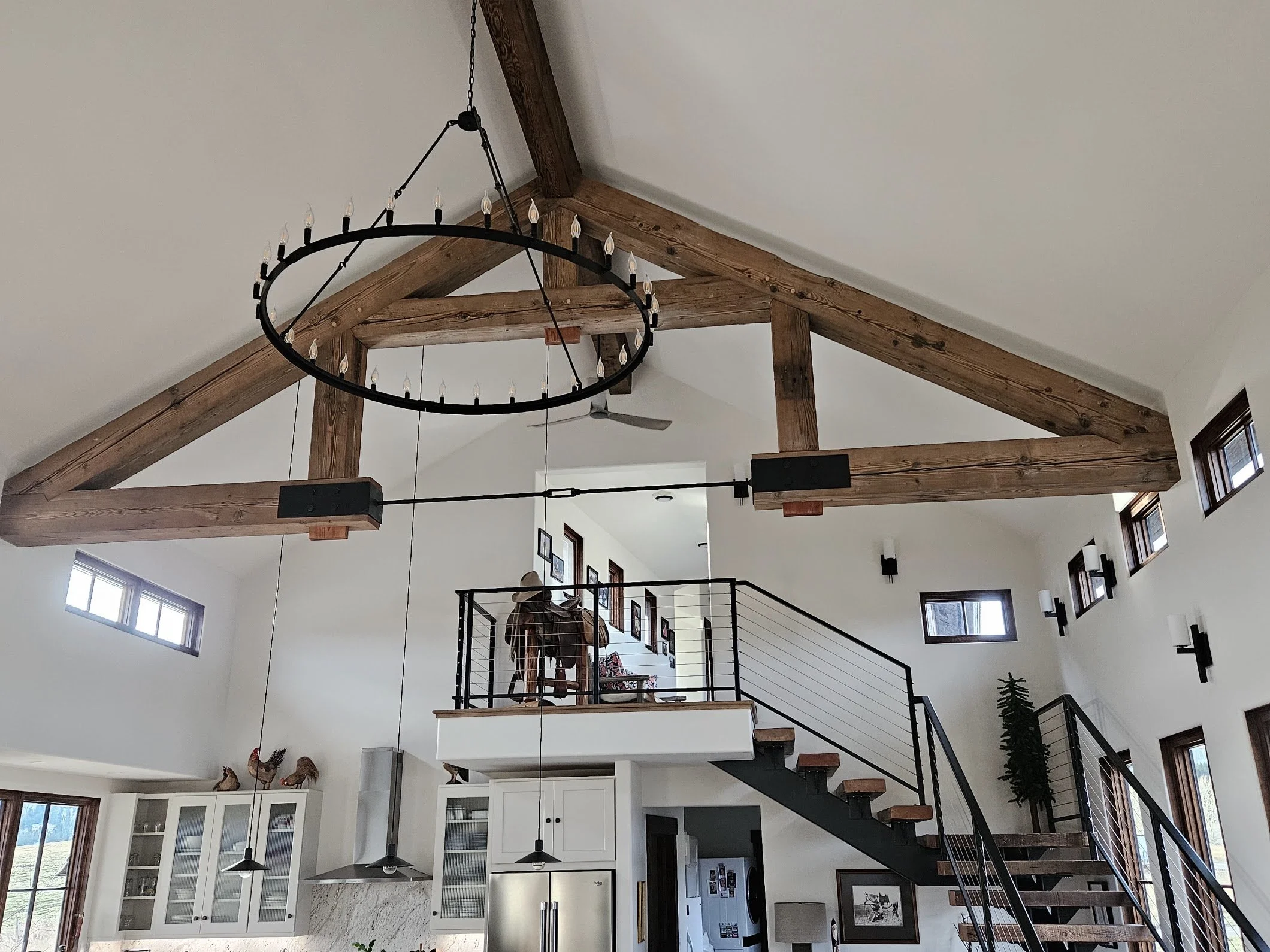 Interior view of a modern home with high vaulted ceiling, exposed wooden beams, a circular chandelier hanging from the ceiling, a staircase with black metal railings, and a mezzanine with a person taking photos. There are multiple small windows providing natural light and decor elements such as framed pictures, a potted plant, and sheep sculptures on top of white cabinets.