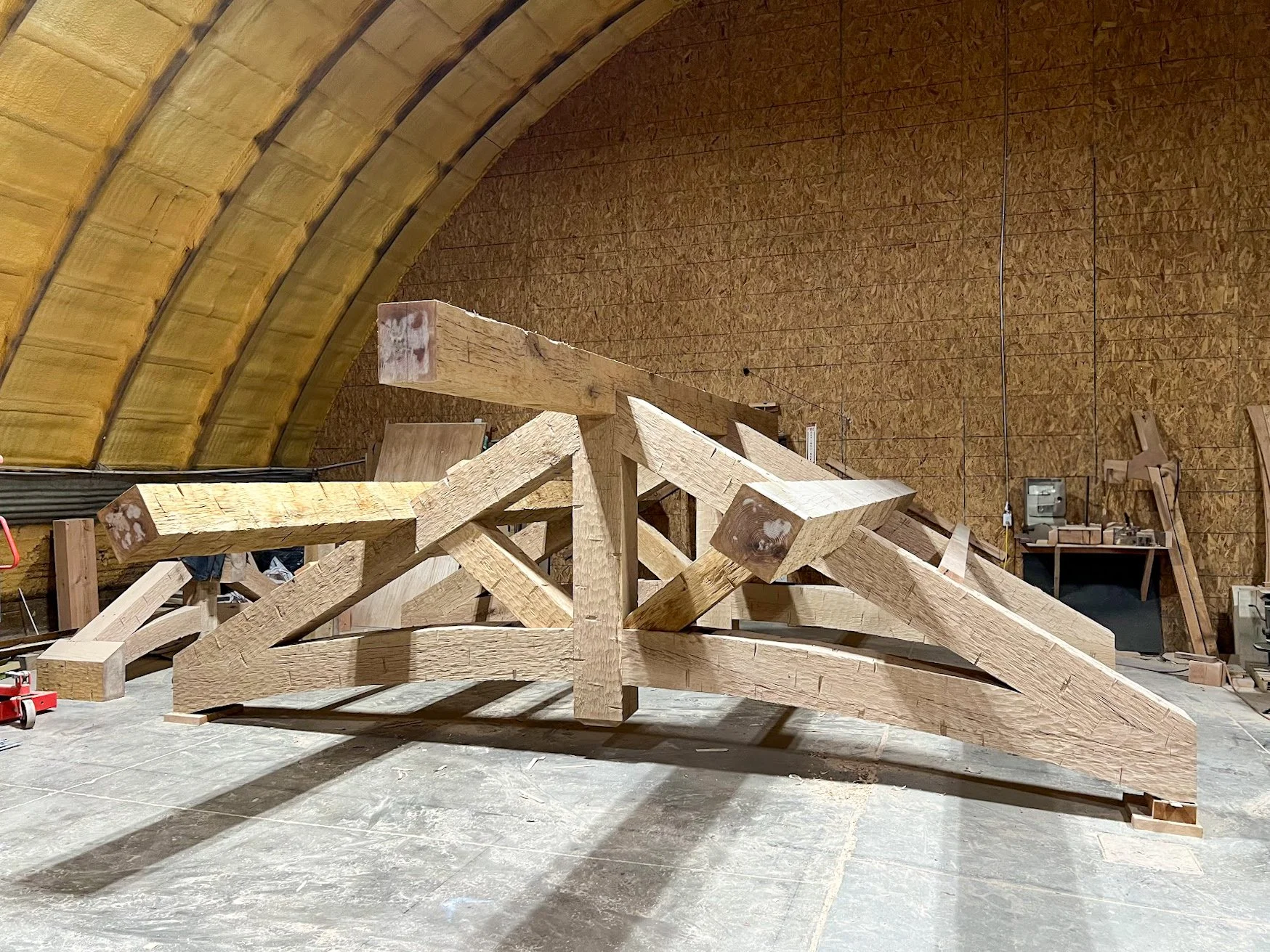 Unfinished wooden structure in a workshop with curved walls and ceiling, tools and equipment in the background.