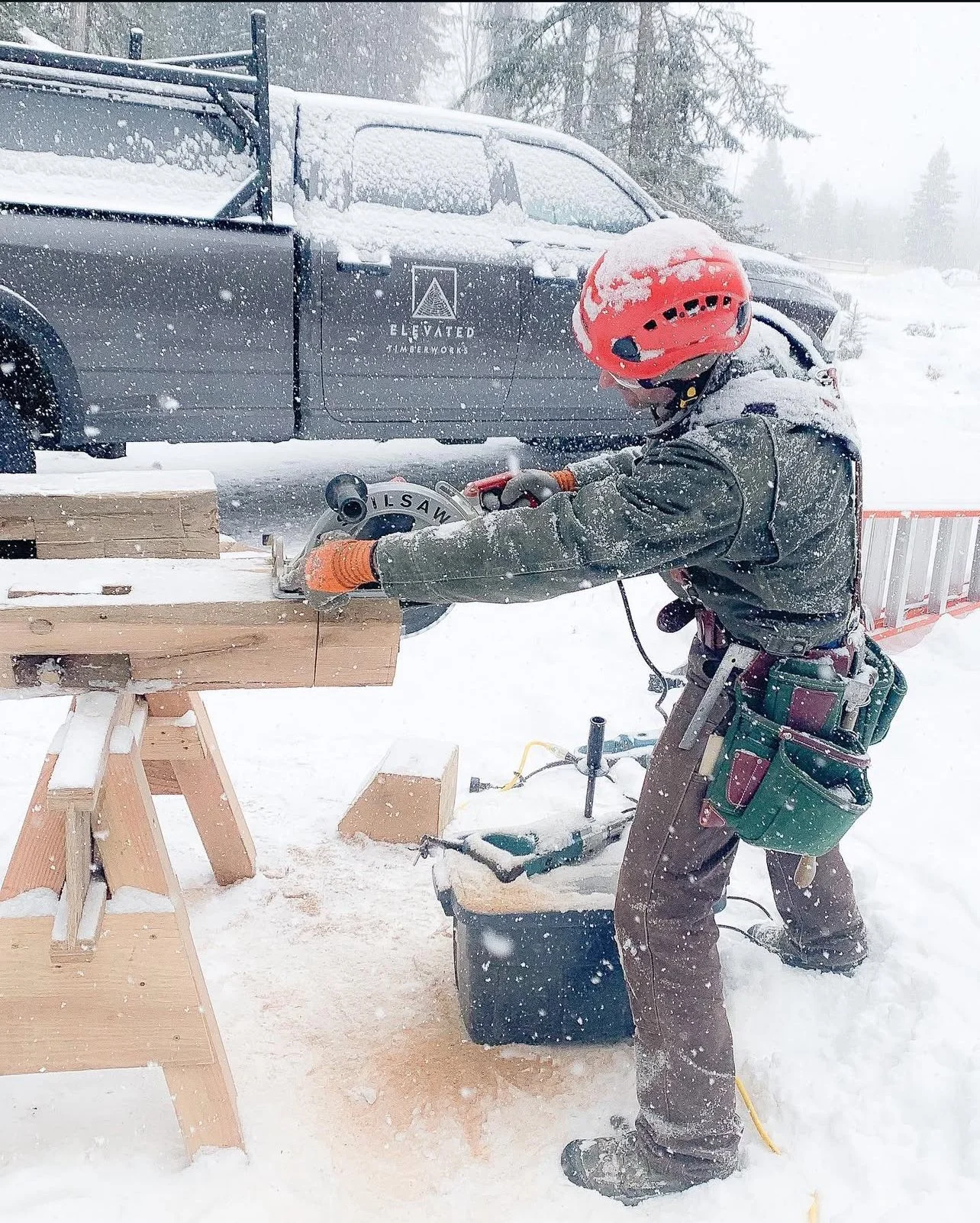 A person wearing a red safety helmet, gray jacket, and brown pants is cutting a wooden beam with a circular saw outdoors in a snowy environment. There is a black vehicle in the background with the logo 'Elevated Timberworks' on the side.