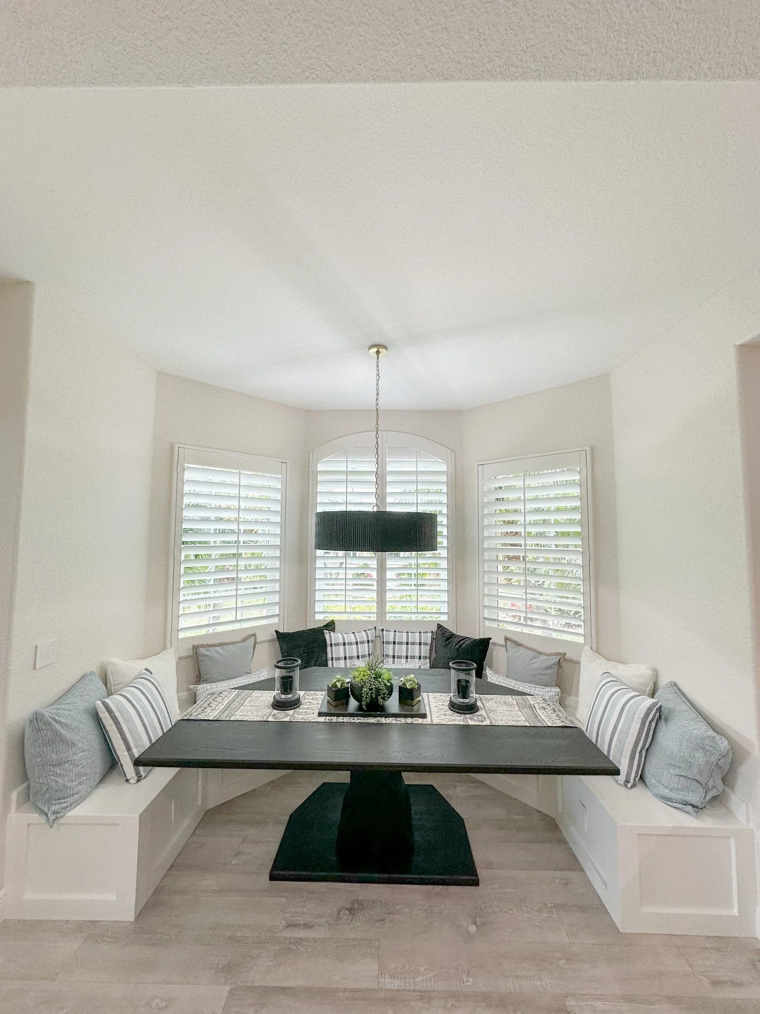 Dining nook with built-in bench seating around a rectangular dark wood table, decorated with a centerpiece and candles, set near three windows with white plantation shutters, and a black drum pendant light hanging above.