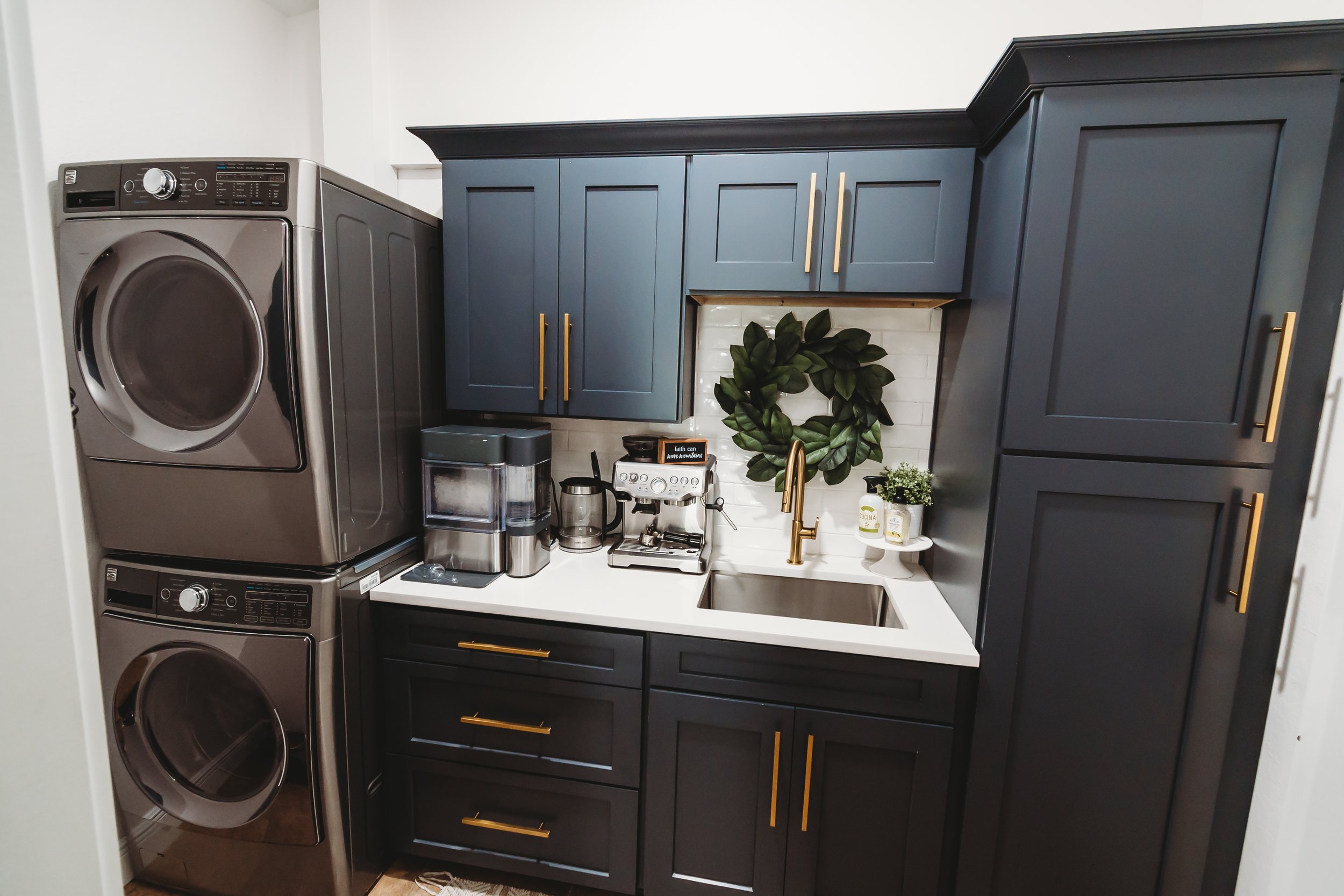 A compact laundry room with navy blue cabinets, gold handles, a white countertop, a small sink with a gold faucet, and stacked metallic washing machine and dryer.
