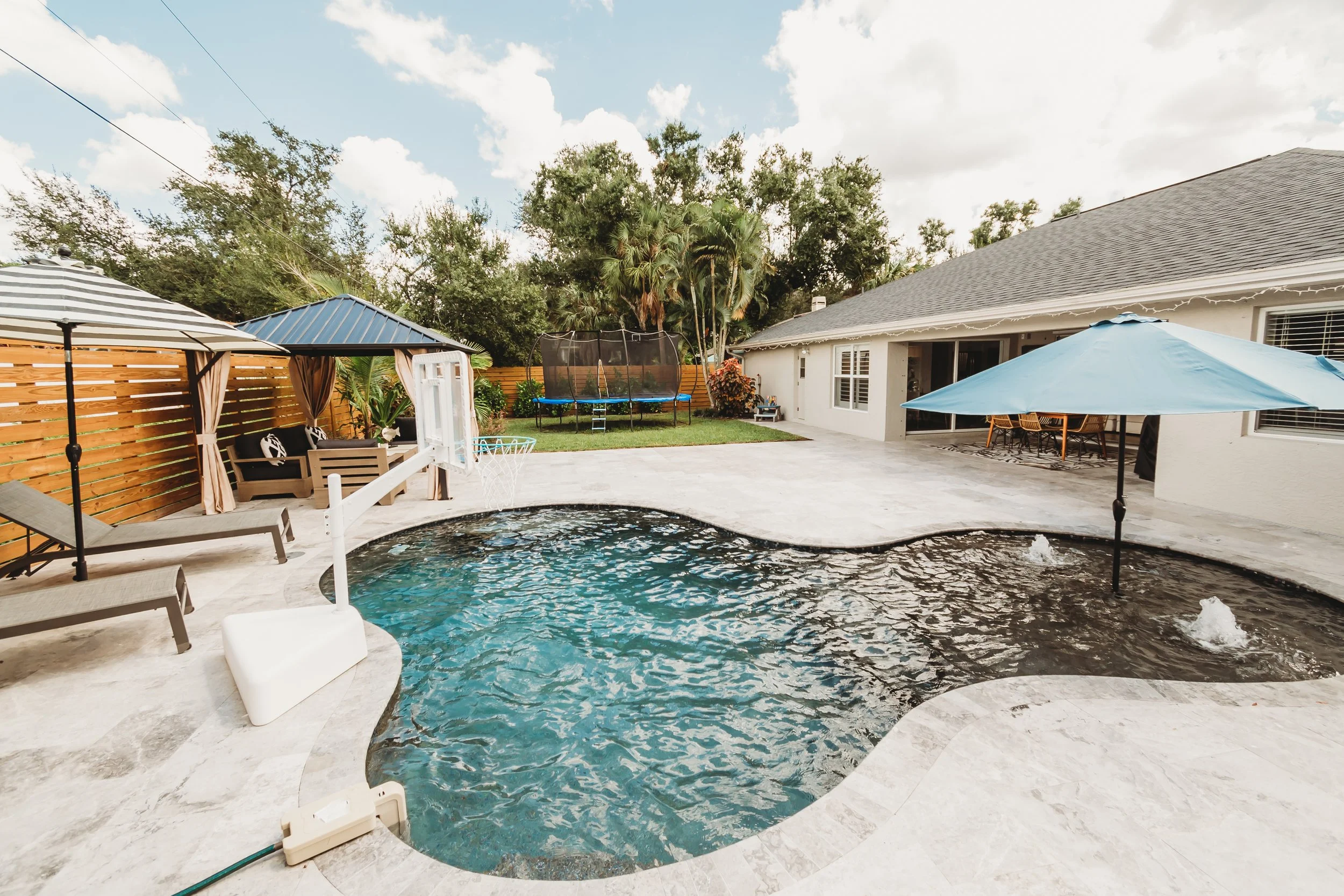 Backyard patio with a swimming pool, shaded seating area, trampoline, and patio table with umbrella.