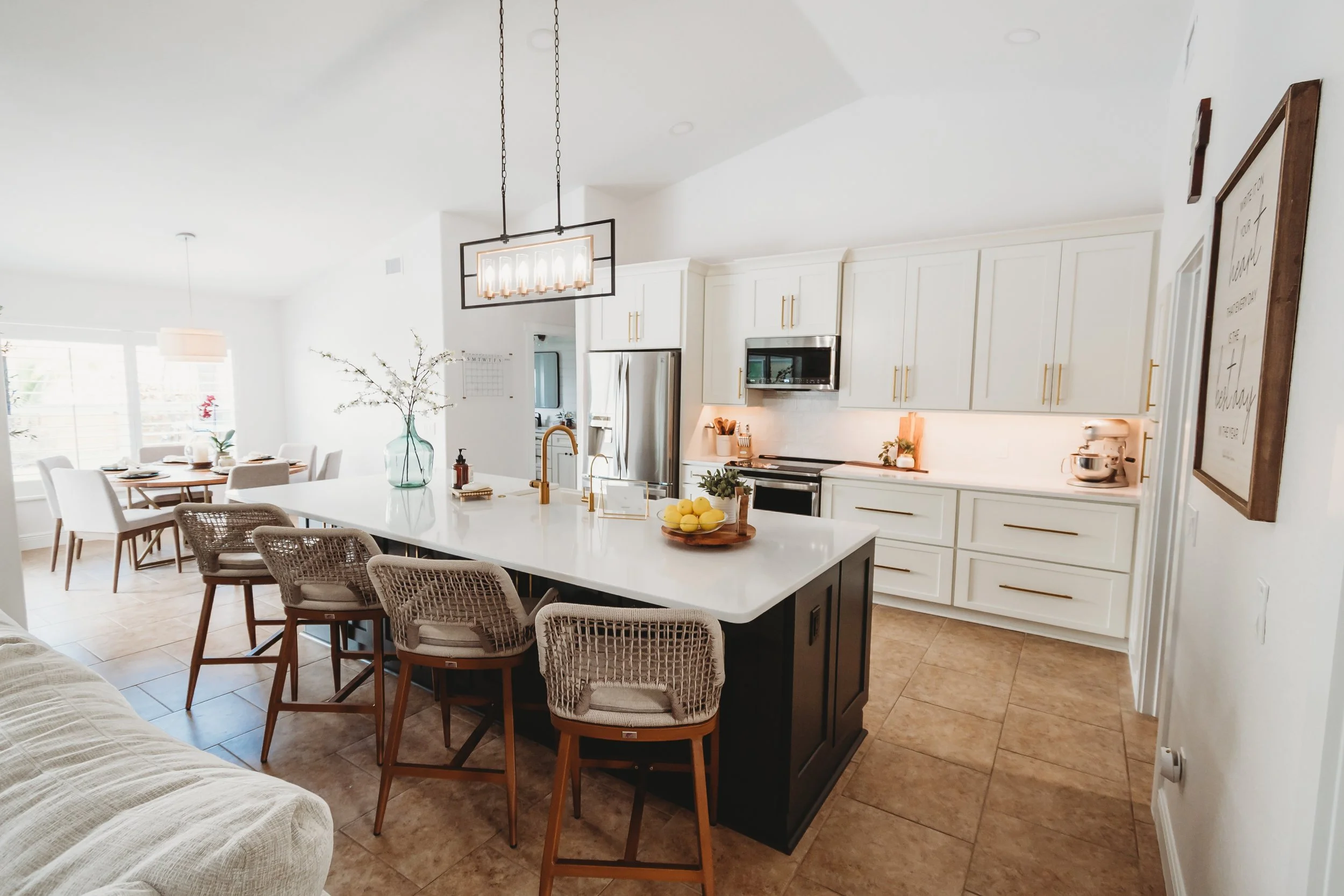 Modern kitchen with white cabinets, black island, beige tile flooring, and a dining area with white chairs in the background.
