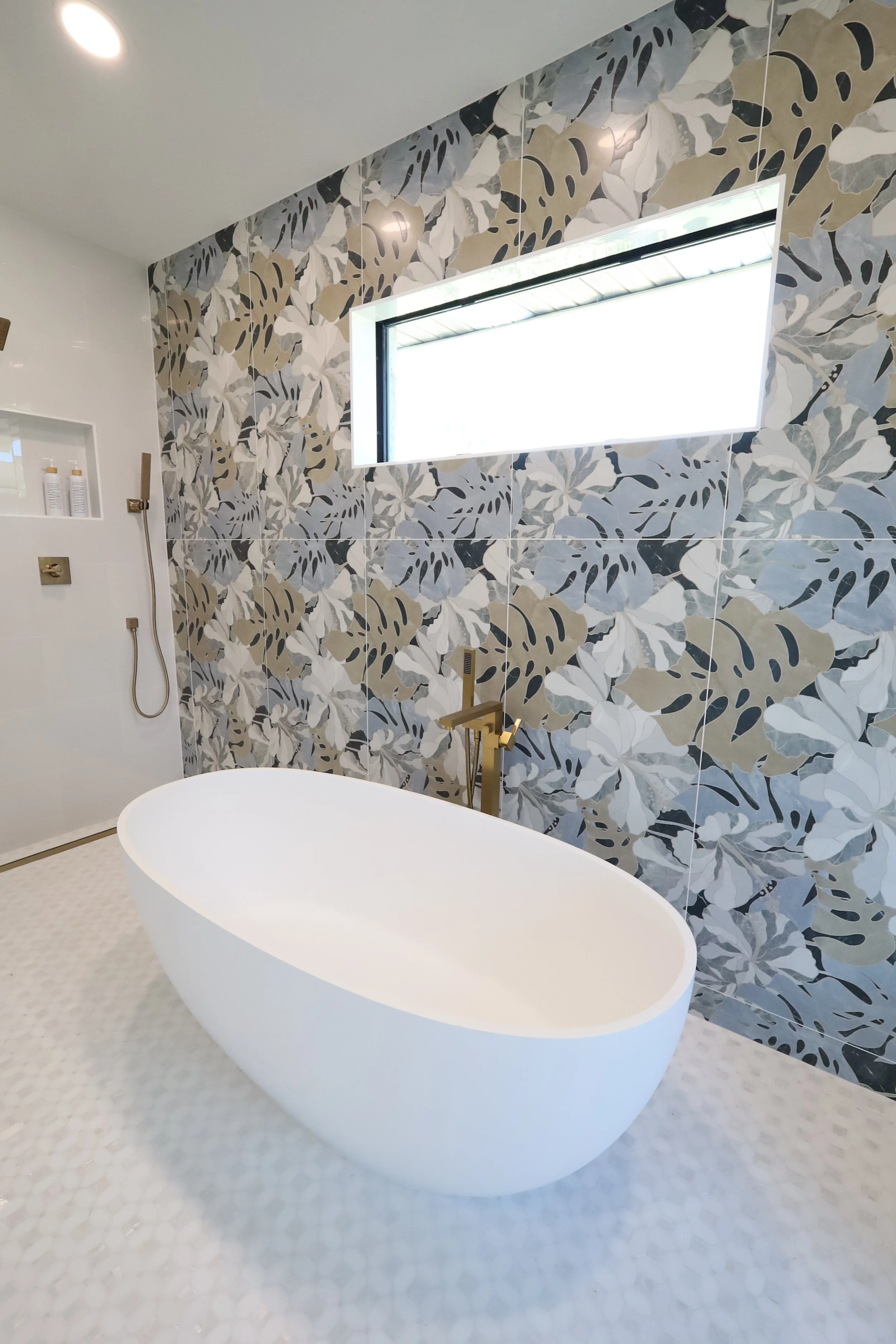 Modern bathroom with a freestanding white bathtub, lush leaf-patterned wall tiles, a rectangular window, and gold fixtures.