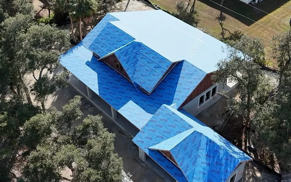 Aerial view of a house under construction with a blue roof partially covered by a protective blue sheeting. Surrounding trees and part of the yard are visible.
