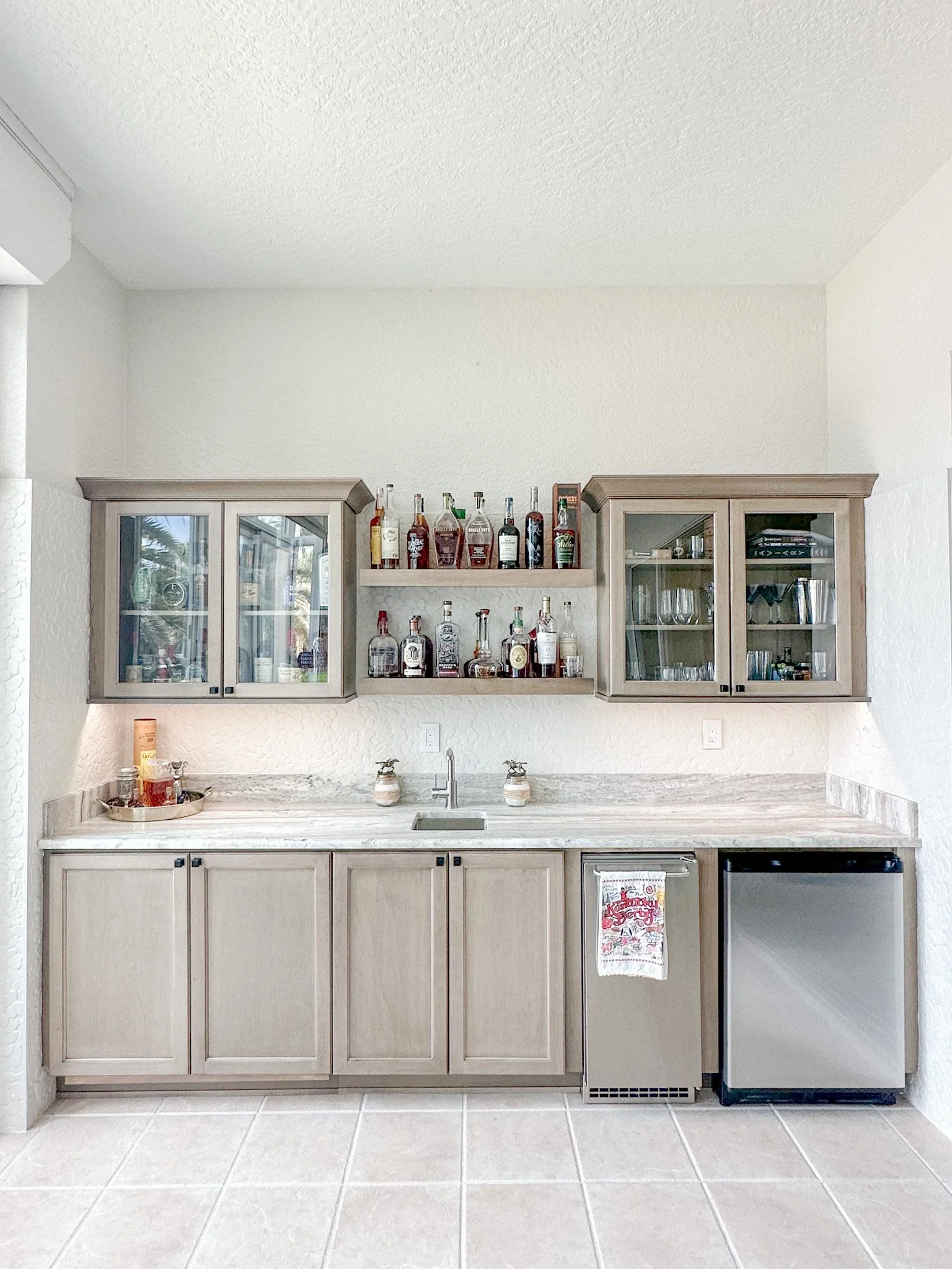 Home bar with upper cabinets, open shelves with liquor bottles, a small fridge, and a marble countertop, featuring a sink and decorative soap dispensers.