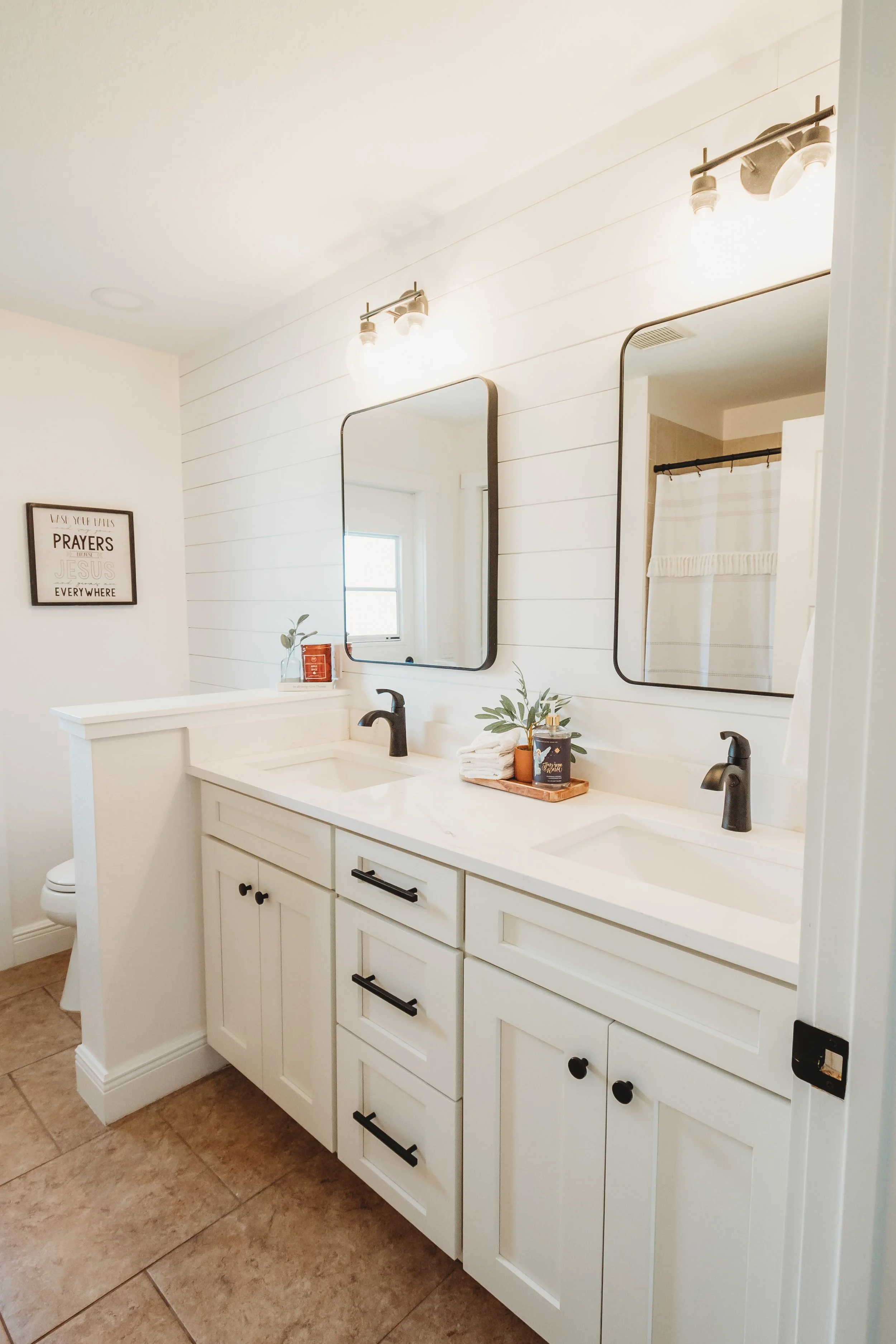 Modern bathroom with a white double vanity, two rectangular mirrors, black faucets, and light fixtures. Decorative items on the counter include plants and toiletries. Tiled floor and a shower with a white curtain are visible.
