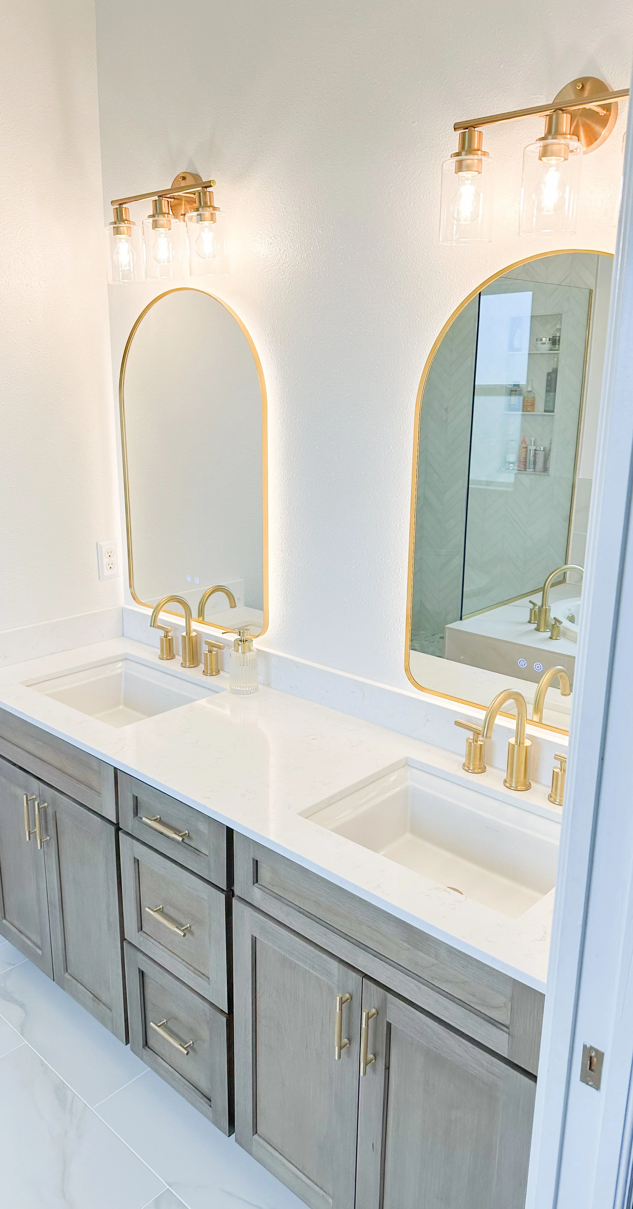 Double bathroom vanity with light wood cabinets, white marble countertop, gold fixtures, two mirrors with gold trim, and wall-mounted light fixtures with exposed bulbs.