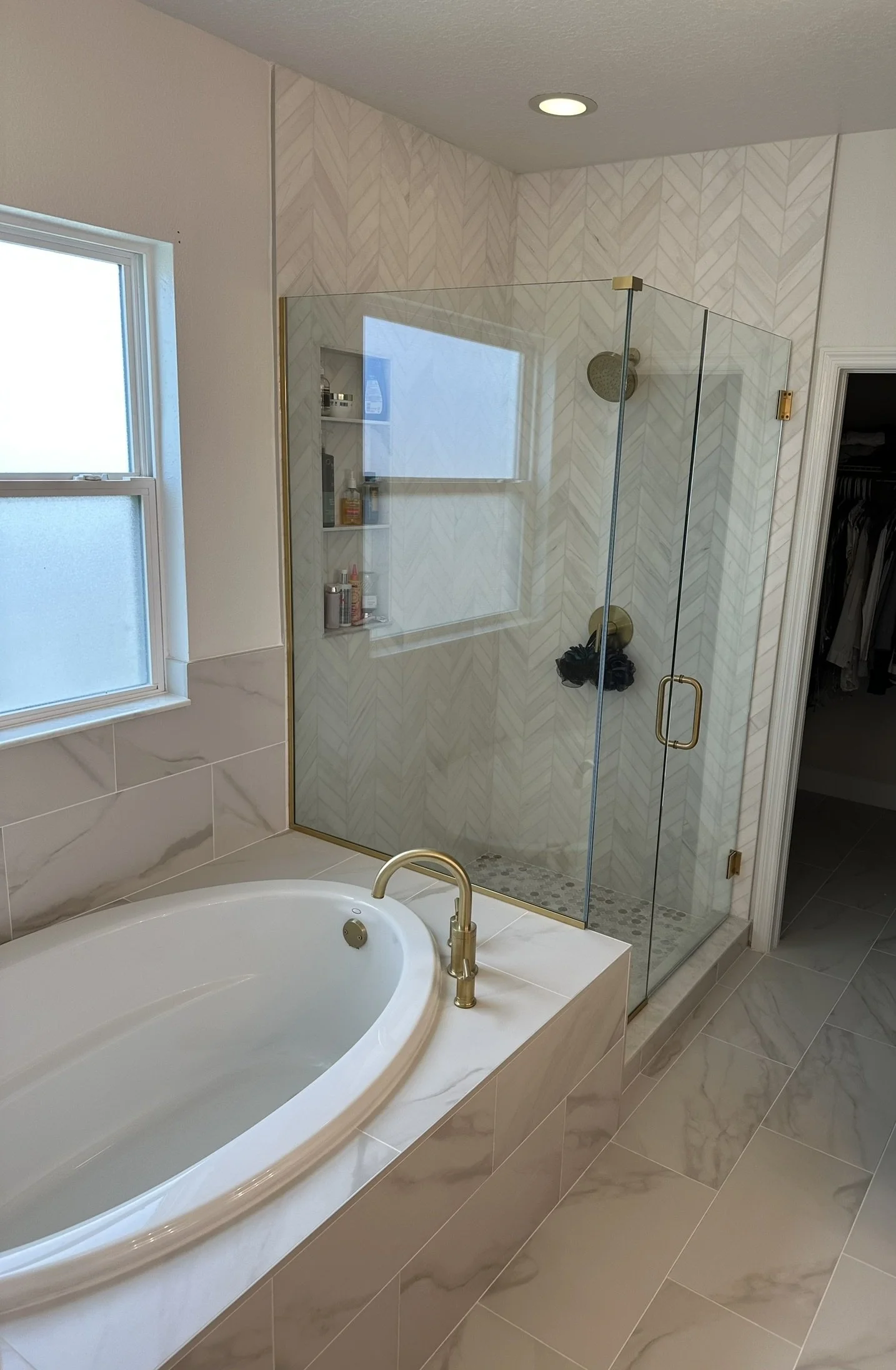 A bathroom featuring a bathtub with a gold faucet, a glass-enclosed shower with a gold handle and fixtures, white marble tiles, a small window, and a walk-in closet visible through an open door.