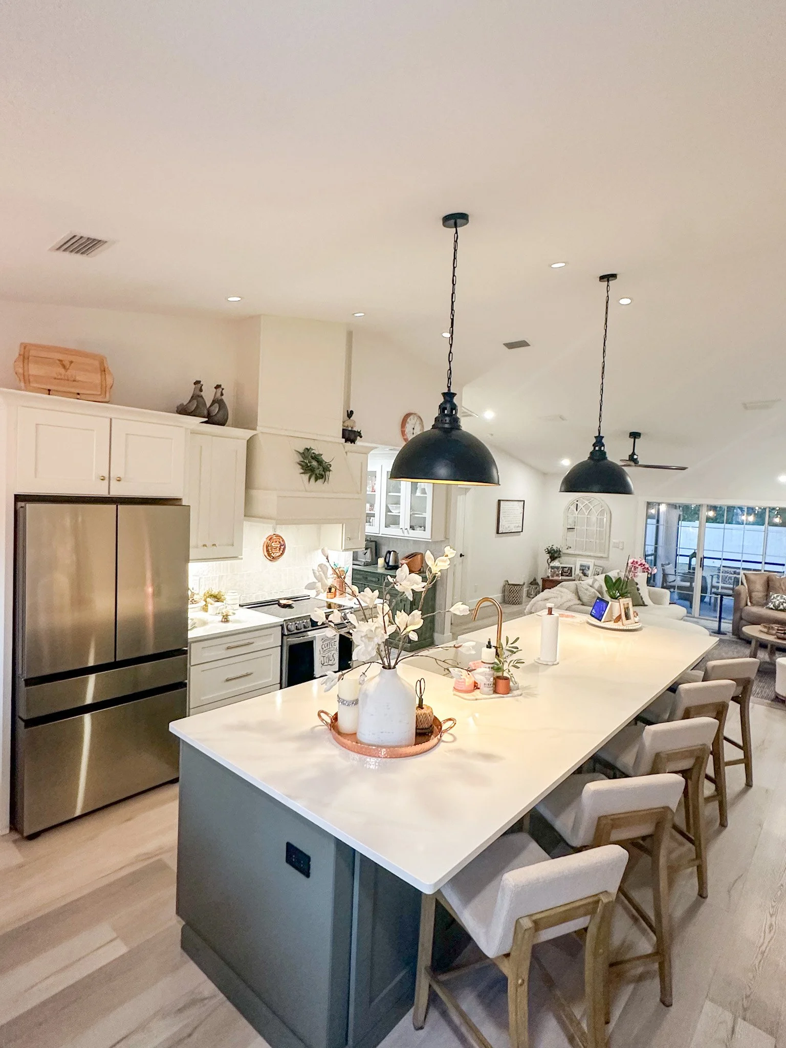 Bright open-concept kitchen with large white island, modern dining chairs, black pendant lights, stainless steel refrigerator, and decorative plants.