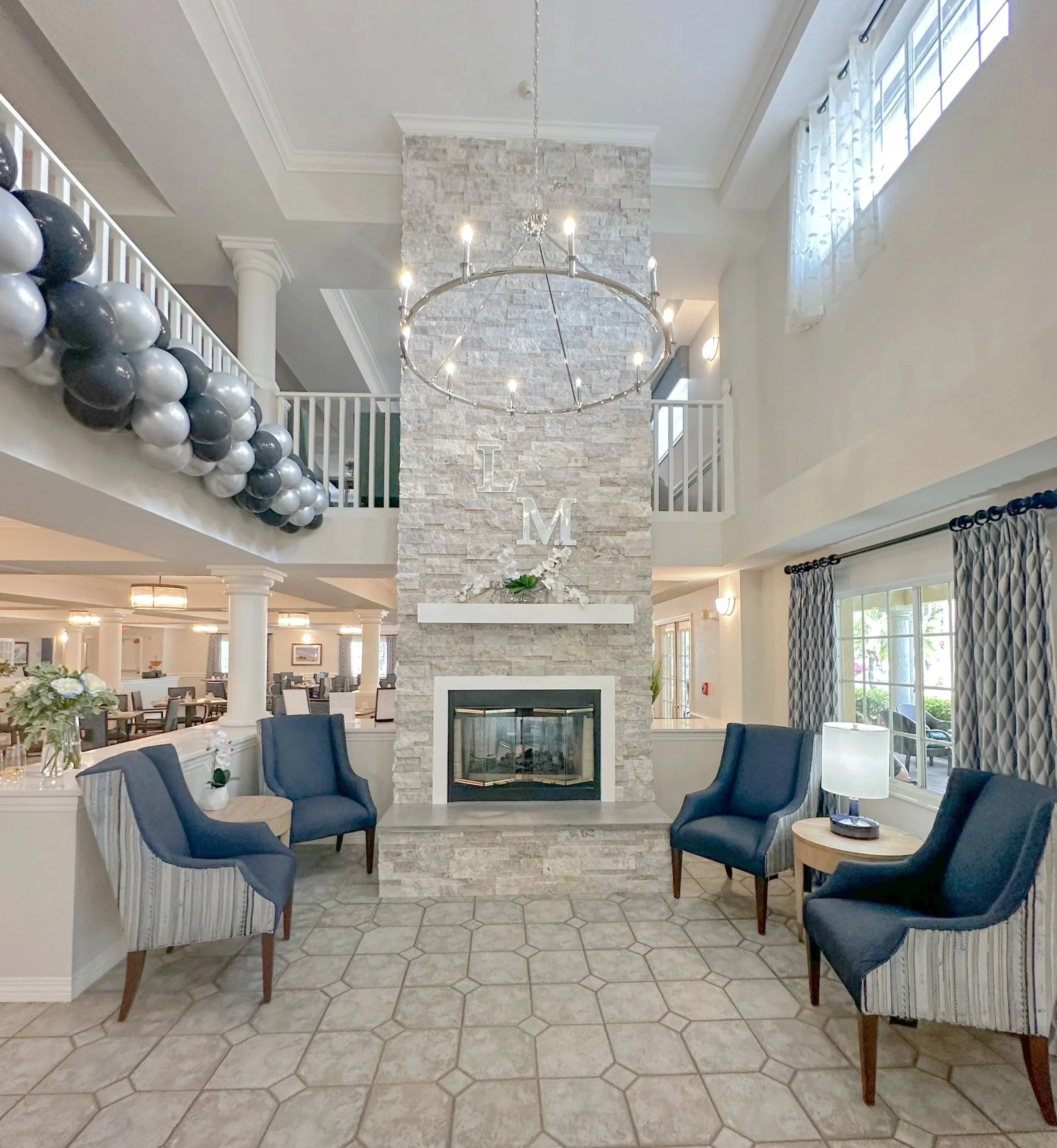 lobby with a stone fireplace, four blue armchairs, a small round table with a lamp, and balloon decorations in shades of black, silver, and gray.