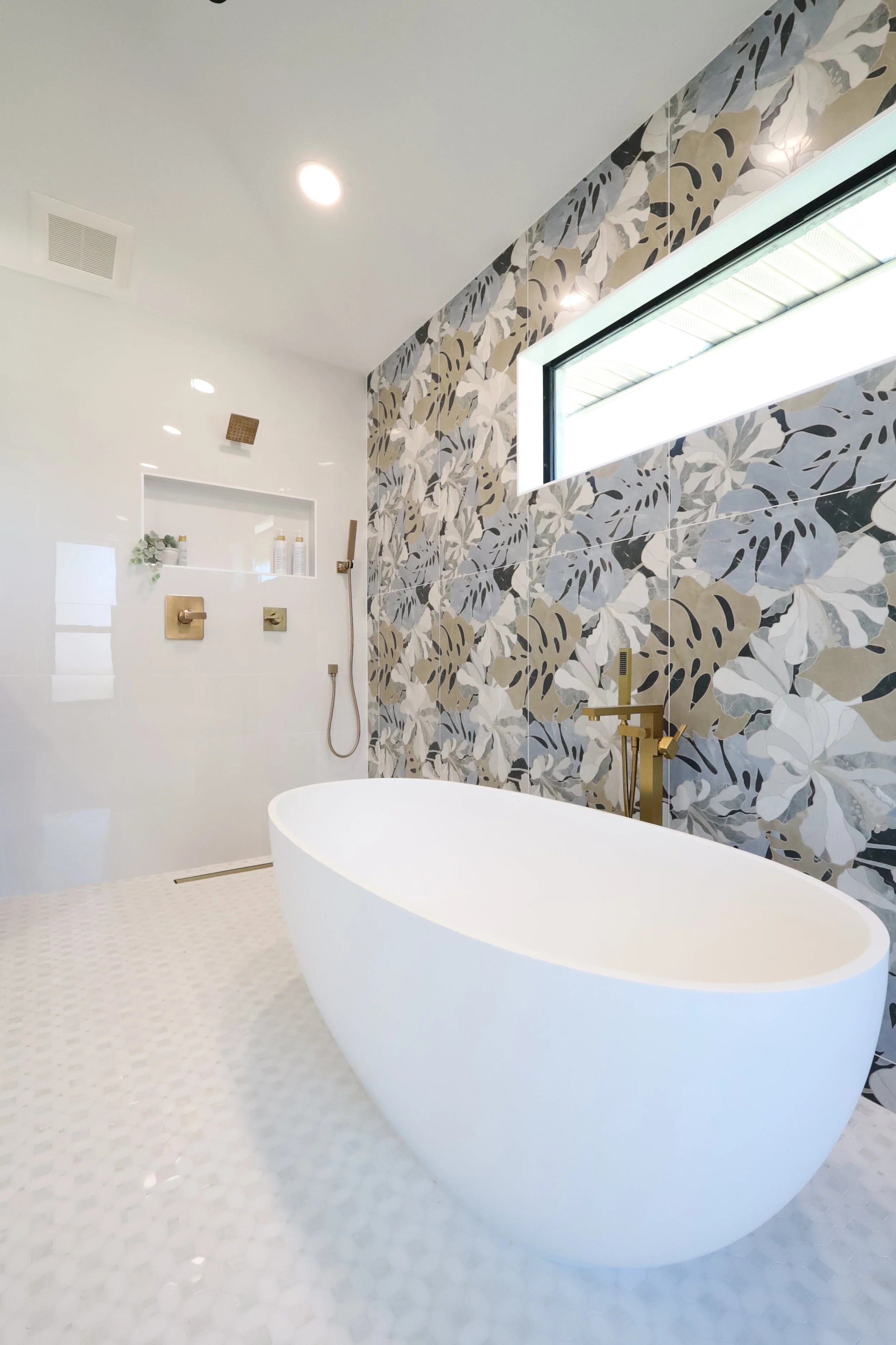 Modern bathroom with a white freestanding bathtub, a wall with leafy patterned tiles, a horizontal window, and a shower area with gold fixtures and a handheld showerhead.