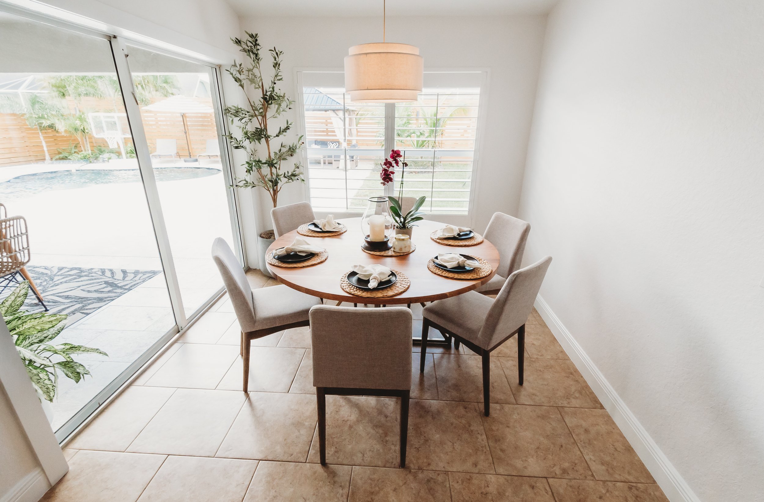 Dining room with a round wooden table set for six, slipcovered chairs, a large window with horizontal blinds, a sliding glass door with view of a backyard pool, and a potted plant.