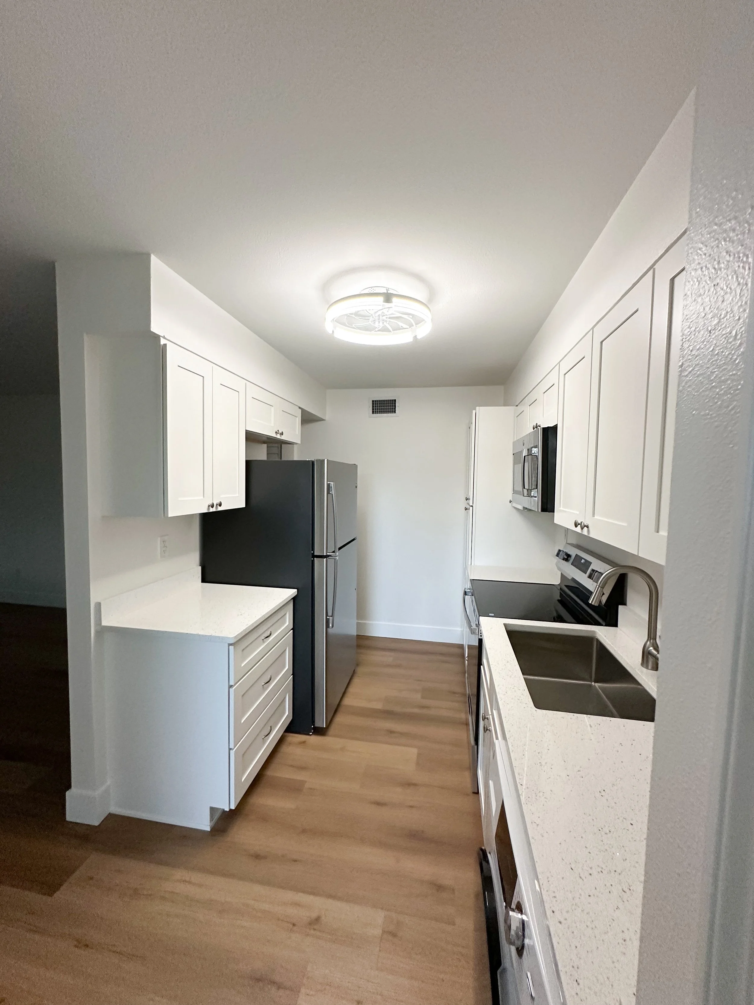 A modern white kitchen with wood flooring, black appliances including a refrigerator, microwave, and stove, white cabinets, a double sink, and a ceiling light fixture.