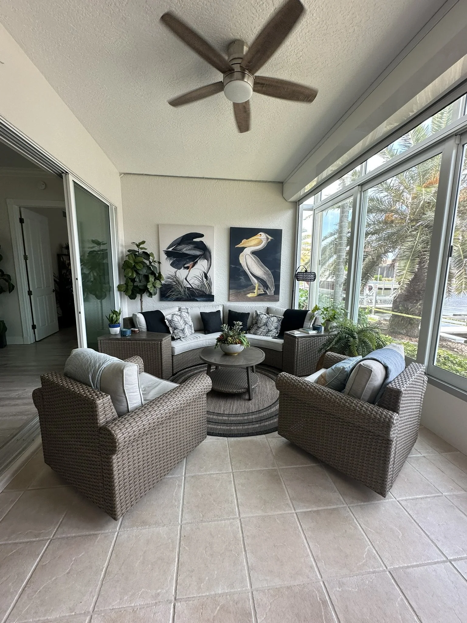 A sunroom with wicker furniture, including a sectional sofa and two armchairs, arranged around a round coffee table with a plant. Large glass windows reveal palm trees outside. Artwork of birds hangs on the wall, and there are potted plants inside.