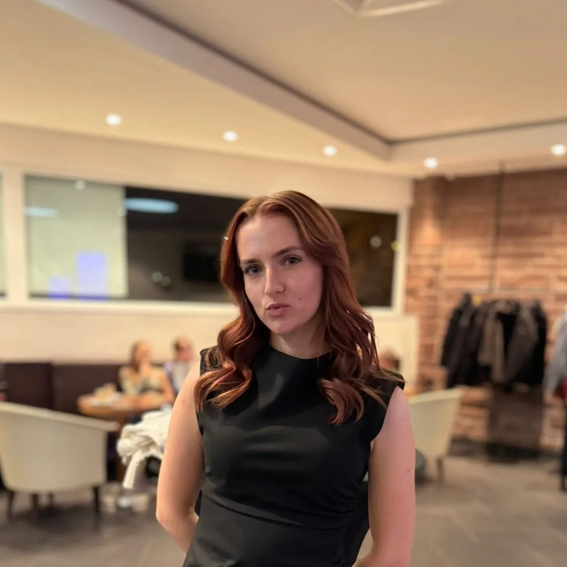 A woman with red hair in loose waves, wearing a sleeveless black dress, standing in a restaurant or cafe with a brick wall, dining tables, and people sitting in the background.