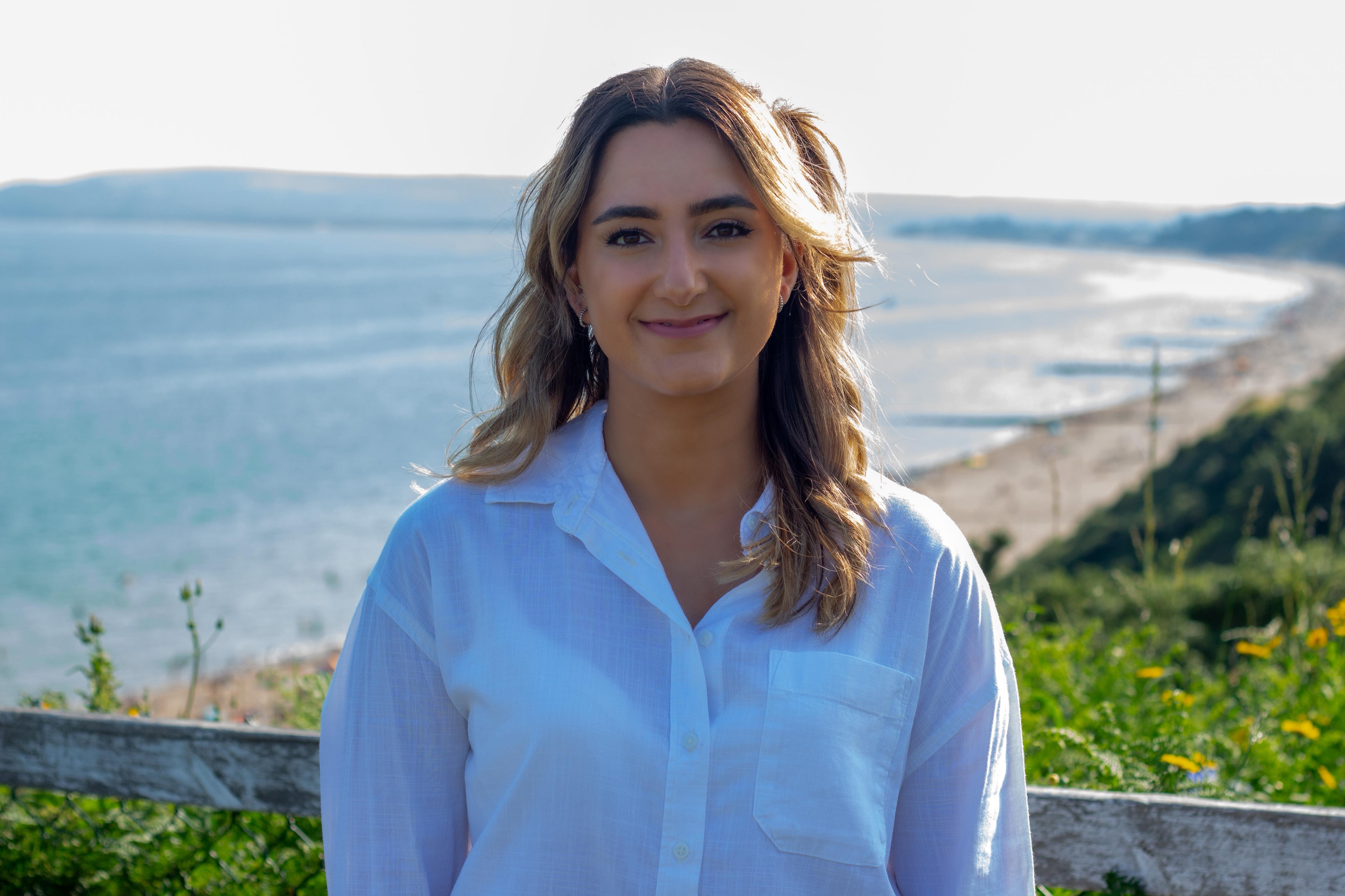 A woman with light brown, wavy hair and dark eyebrows stands by the coast with the ocean and cliffs in the background, smiling gently at the camera.