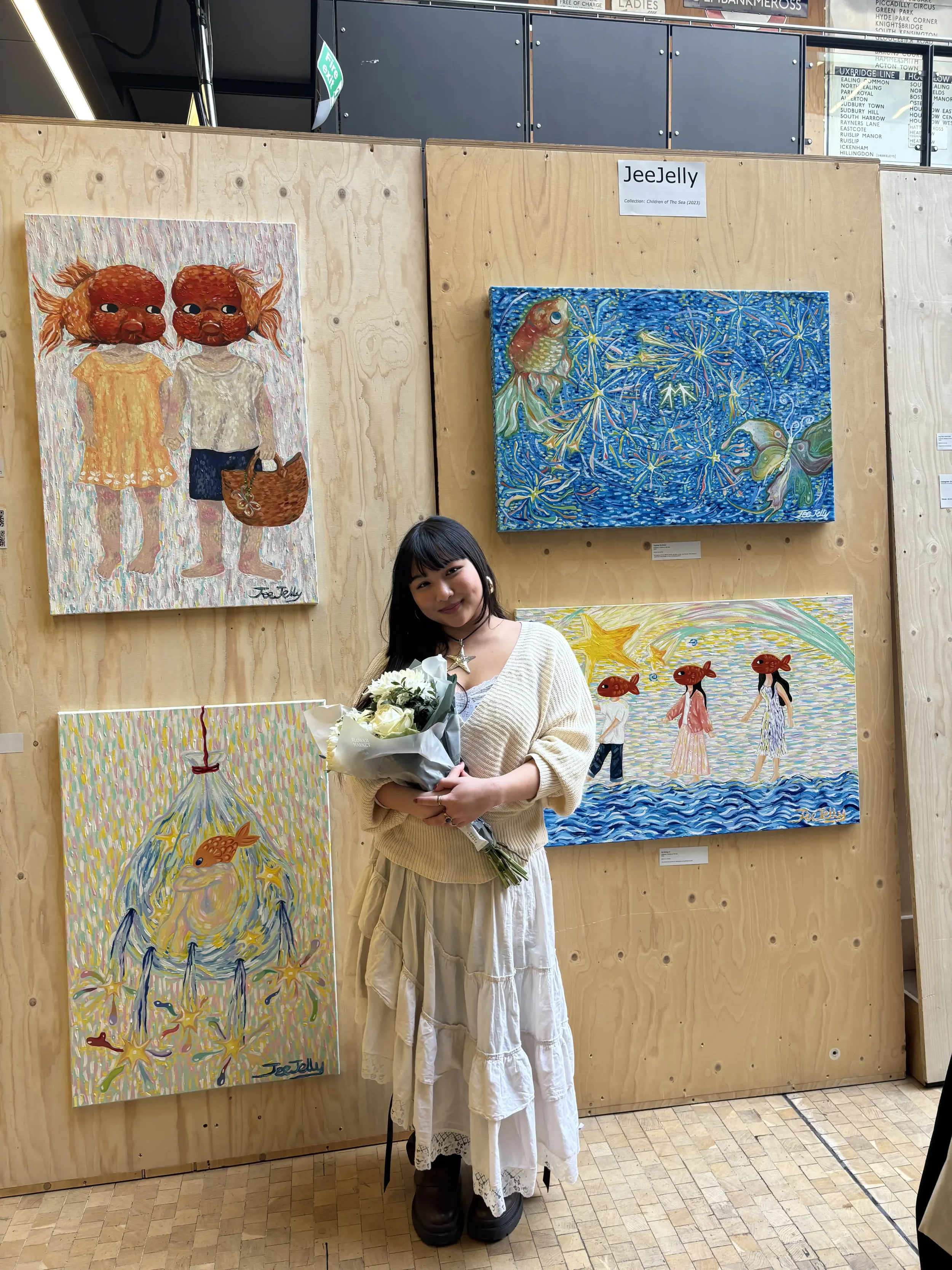A woman with dark hair, wearing a cream-colored sweater and a white skirt, holding a bouquet of white flowers, standing in front of artwork that depicts whimsical fish with human-like heads and a child with a fish head, in an art gallery with wooden wall panels.