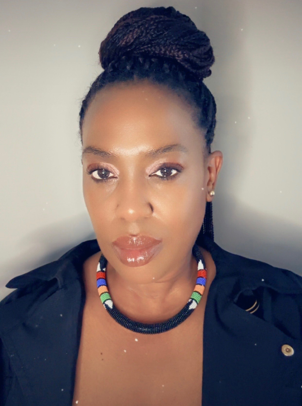 A woman with dark skin and braided hair styled in an updo, wearing a black top and a beaded necklace with colorful beads, looking directly at the camera with a neutral expression.