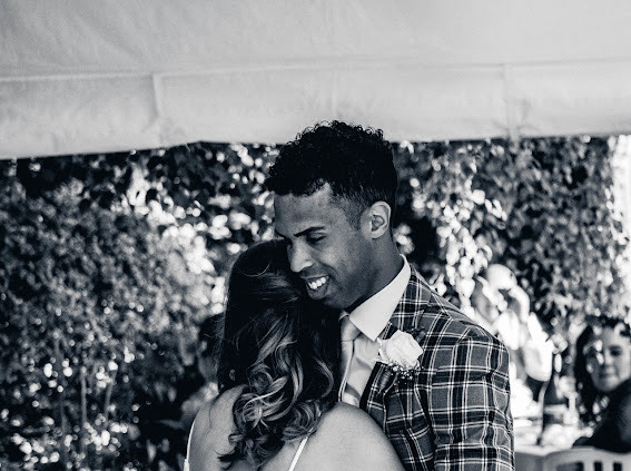 A man and woman embracing at a wedding reception, with trees and other guests in the background.