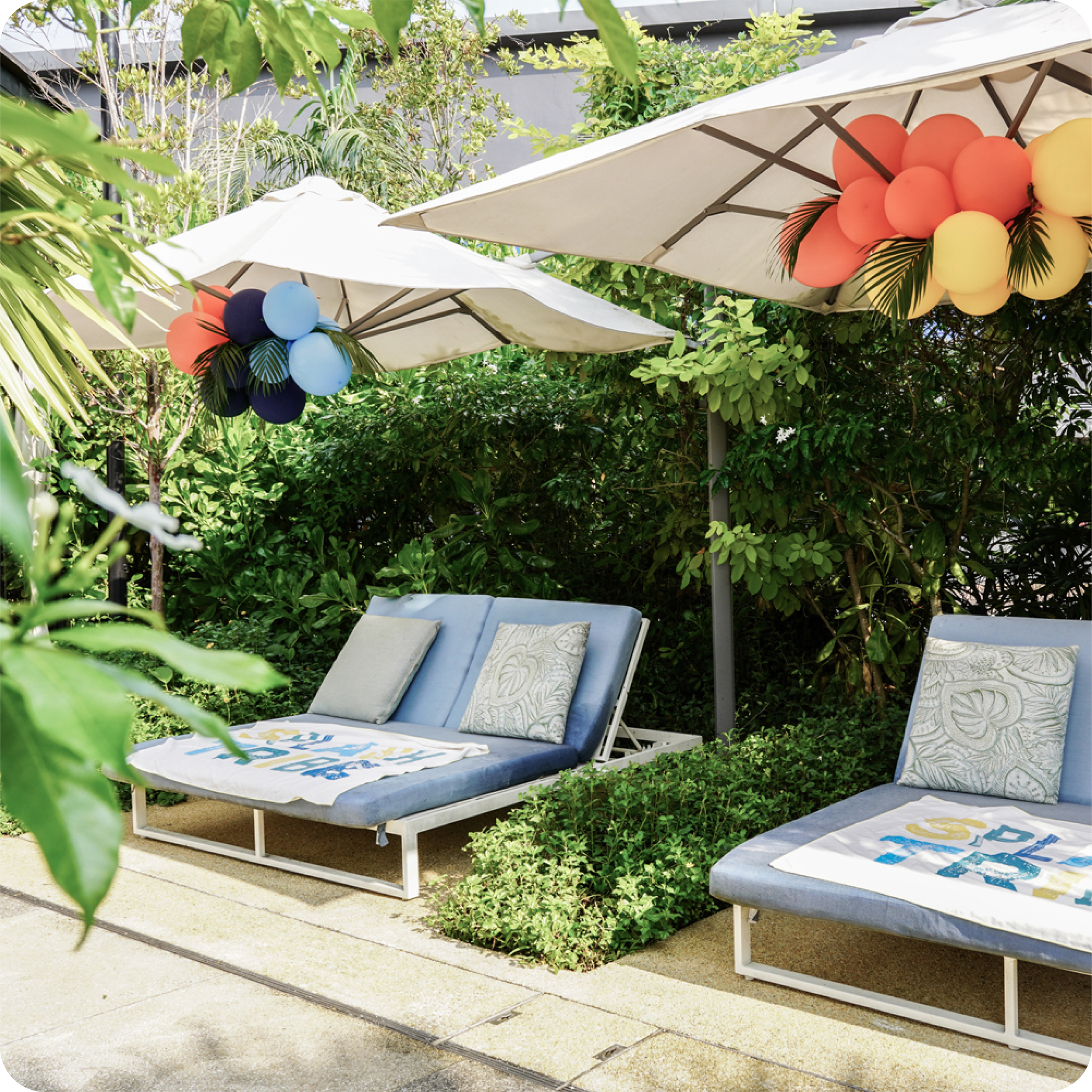 Outdoor patio with two blue lounge chairs under umbrellas decorated with balloons, amid green foliage. Event design by Studio Mojo 