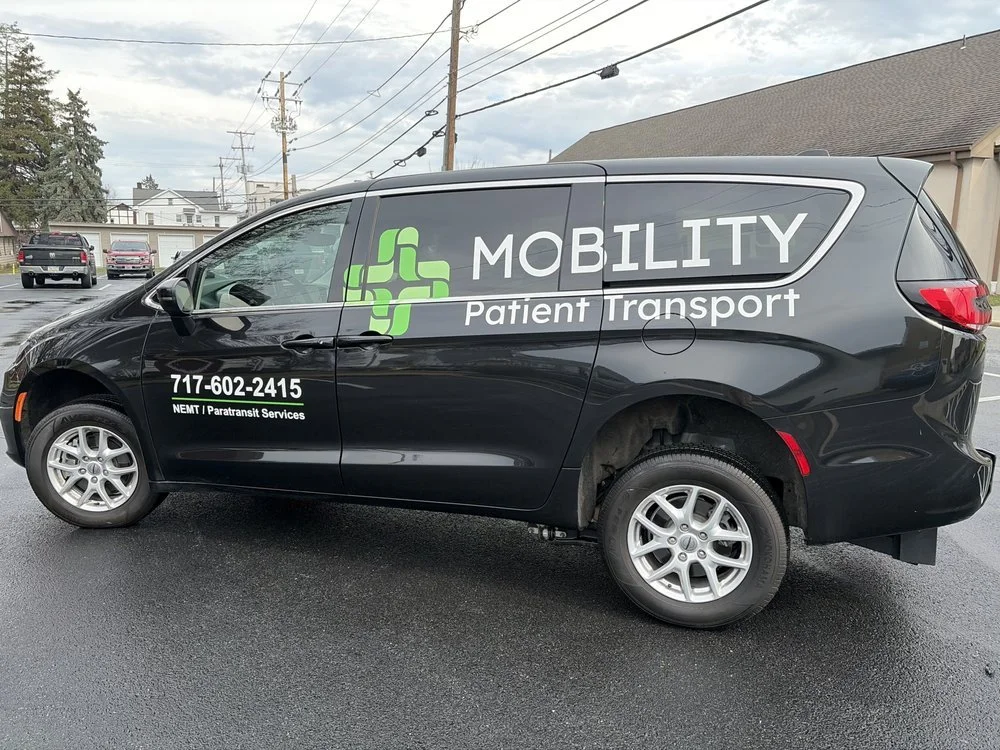 Black vehicle with signage that reads 'Mobility Patient Transport' and a phone number, parked on a wet street with houses and trees in the background.
