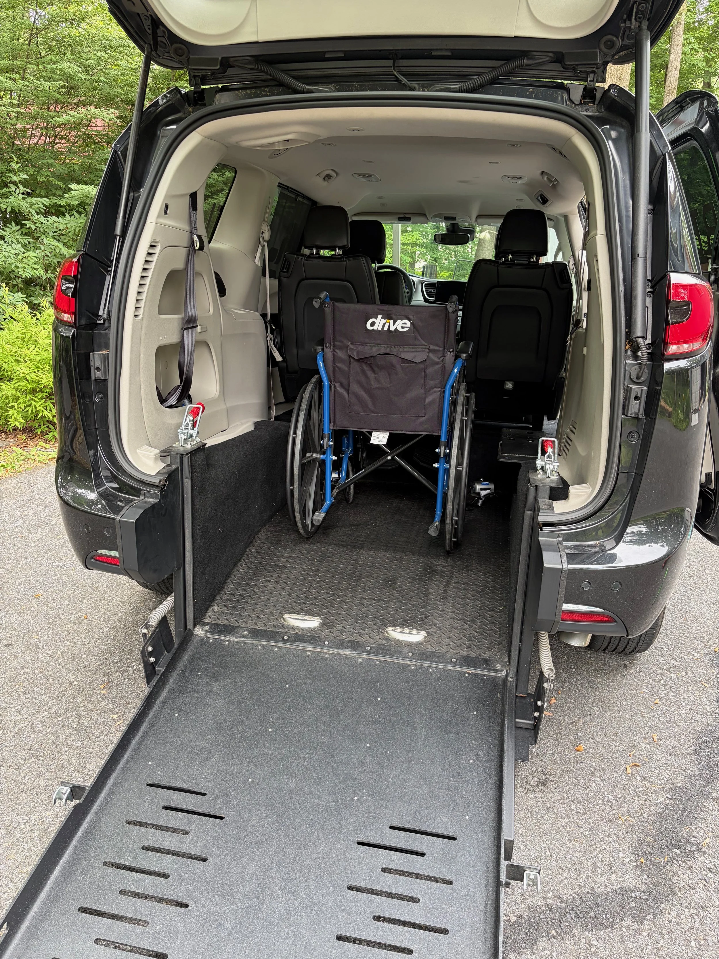 The back of a black van with the rear hatch open, revealing a wheelchair and an empty loading area, surrounded by trees with green leaves.