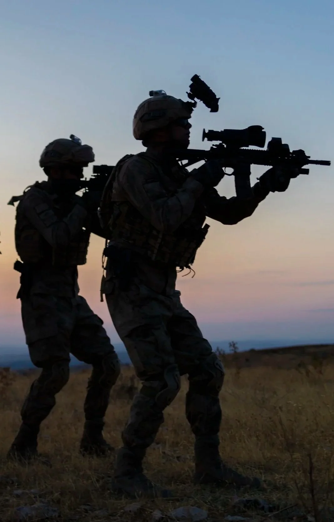 Silhouettes of two soldiers carrying rifles while walking on grassy terrain during sunset or dawn.