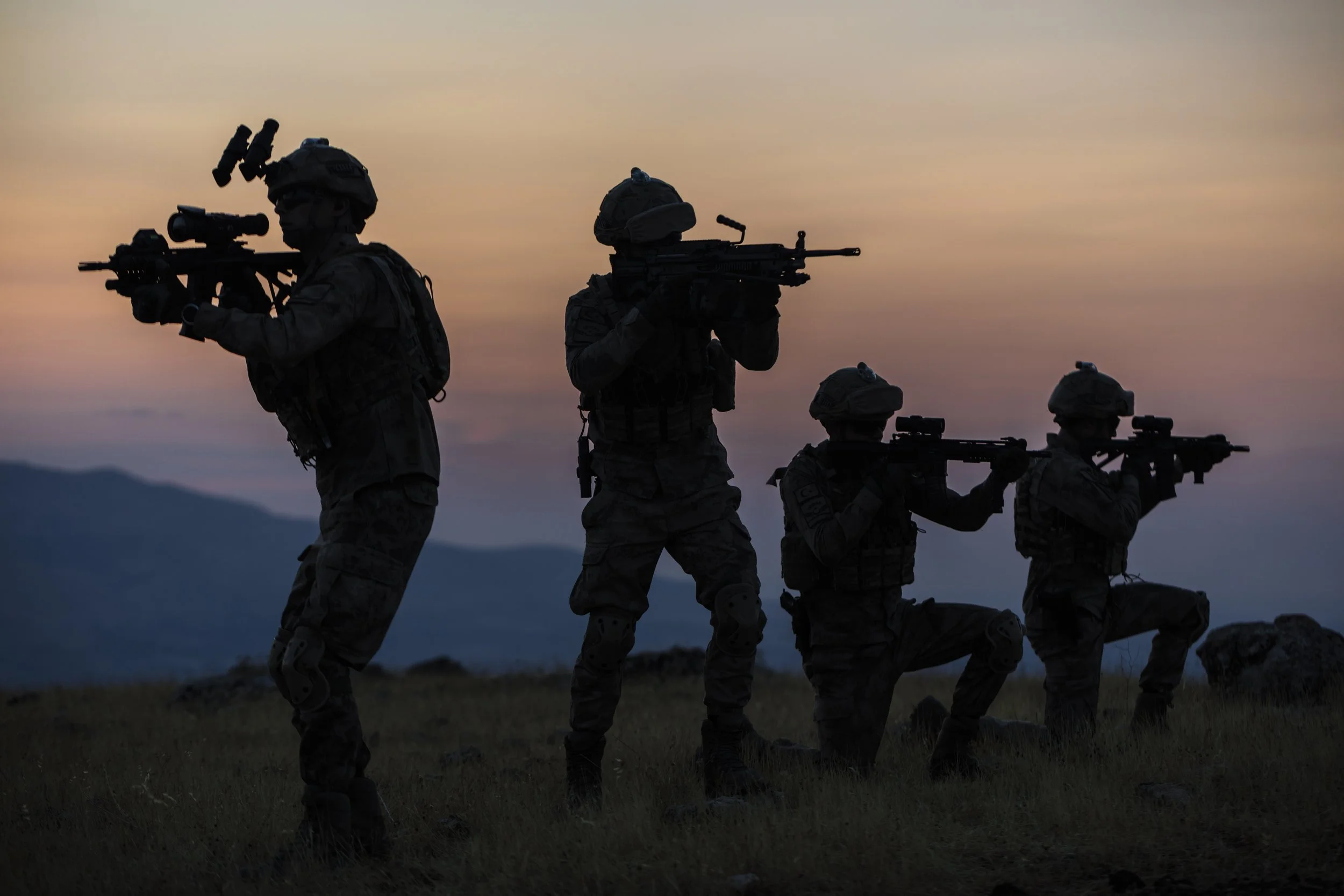 Four soldiers standing in a field at sunset, holding rifles in shooting positions.