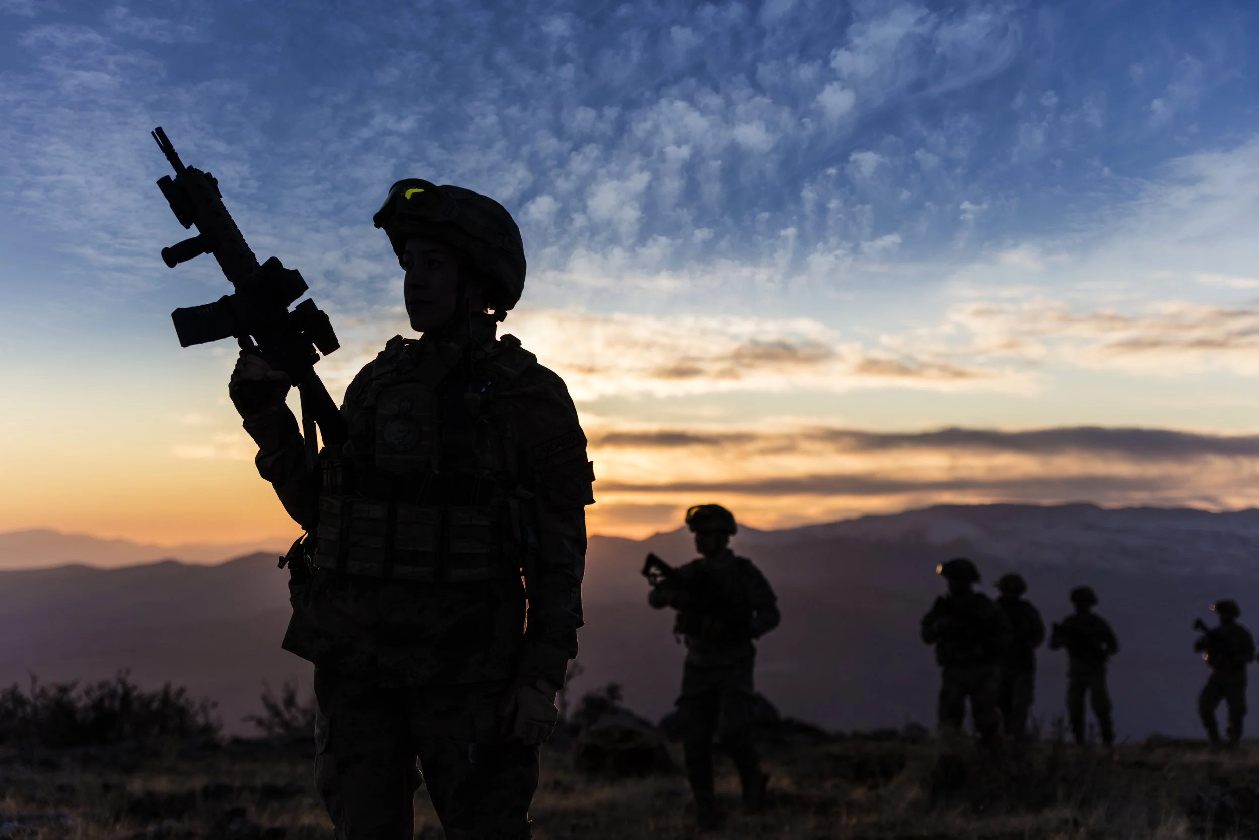 Silhouettes of soldiers in combat gear holding rifles at sunset over a mountain landscape.