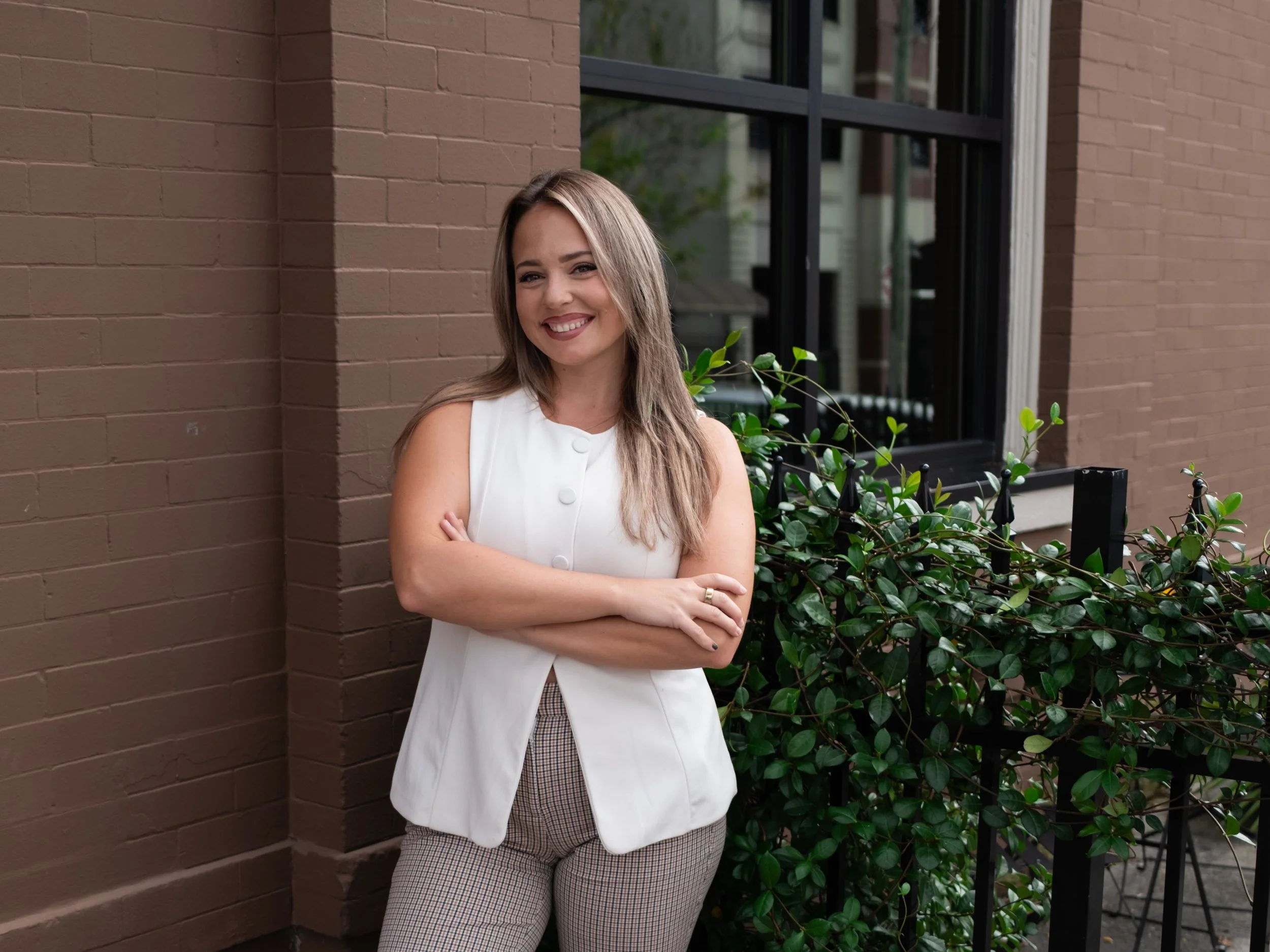 A young woman with long hair wearing a white sleeveless top and checkered pants, smiling with arms crossed, standing outdoors against a brick wall, with greenery and a window in the background.