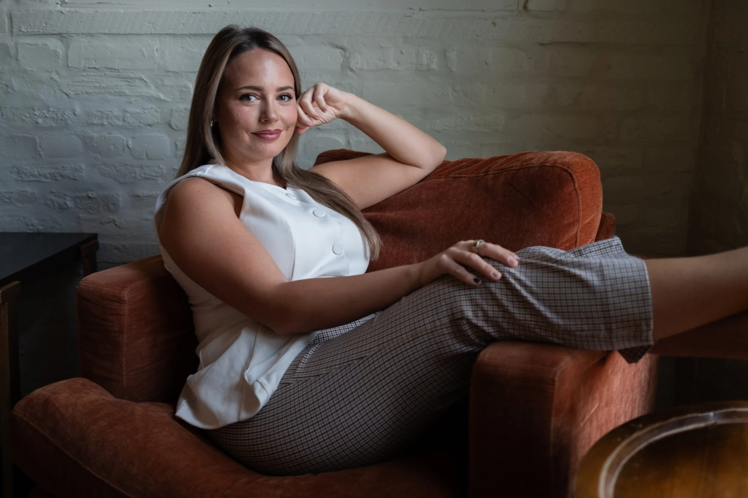 A woman sitting on a burnt orange sofa, resting her head on her hand, smiling, wearing a white sleeveless top and plaid shorts, with a white brick wall in the background.