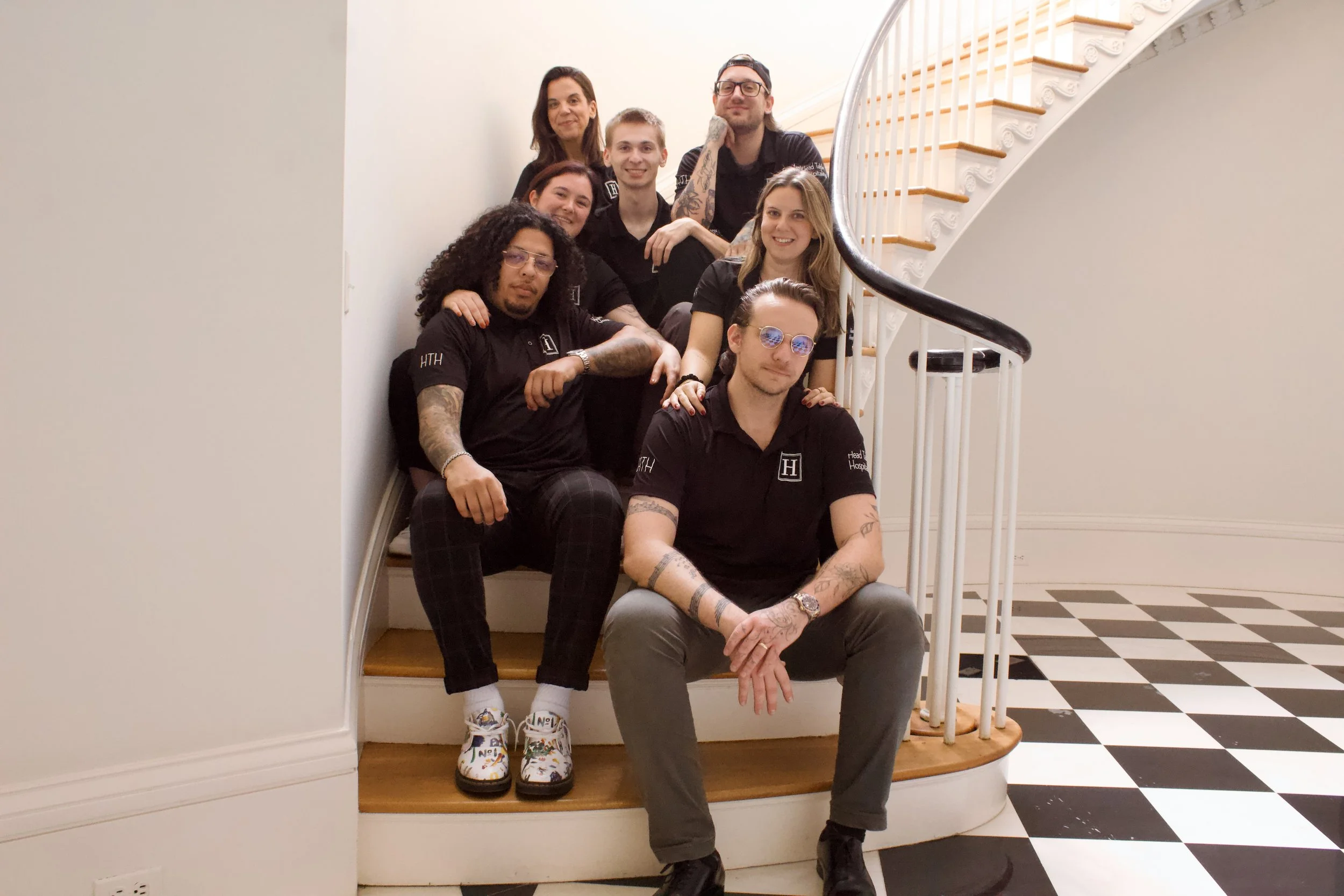 Group of nine young adults sitting on a staircase inside a building with white walls and a black-and-white checkered floor.