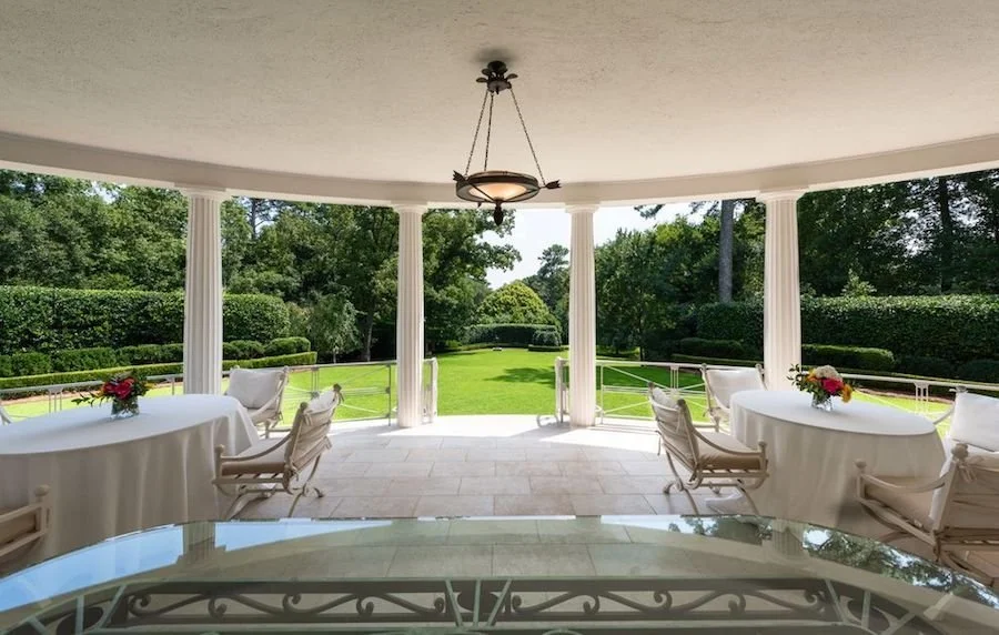 Elegant covered porch with round tables, white chairs, floral centerpieces, and a view of a well-manicured garden with green trees and shrubs.