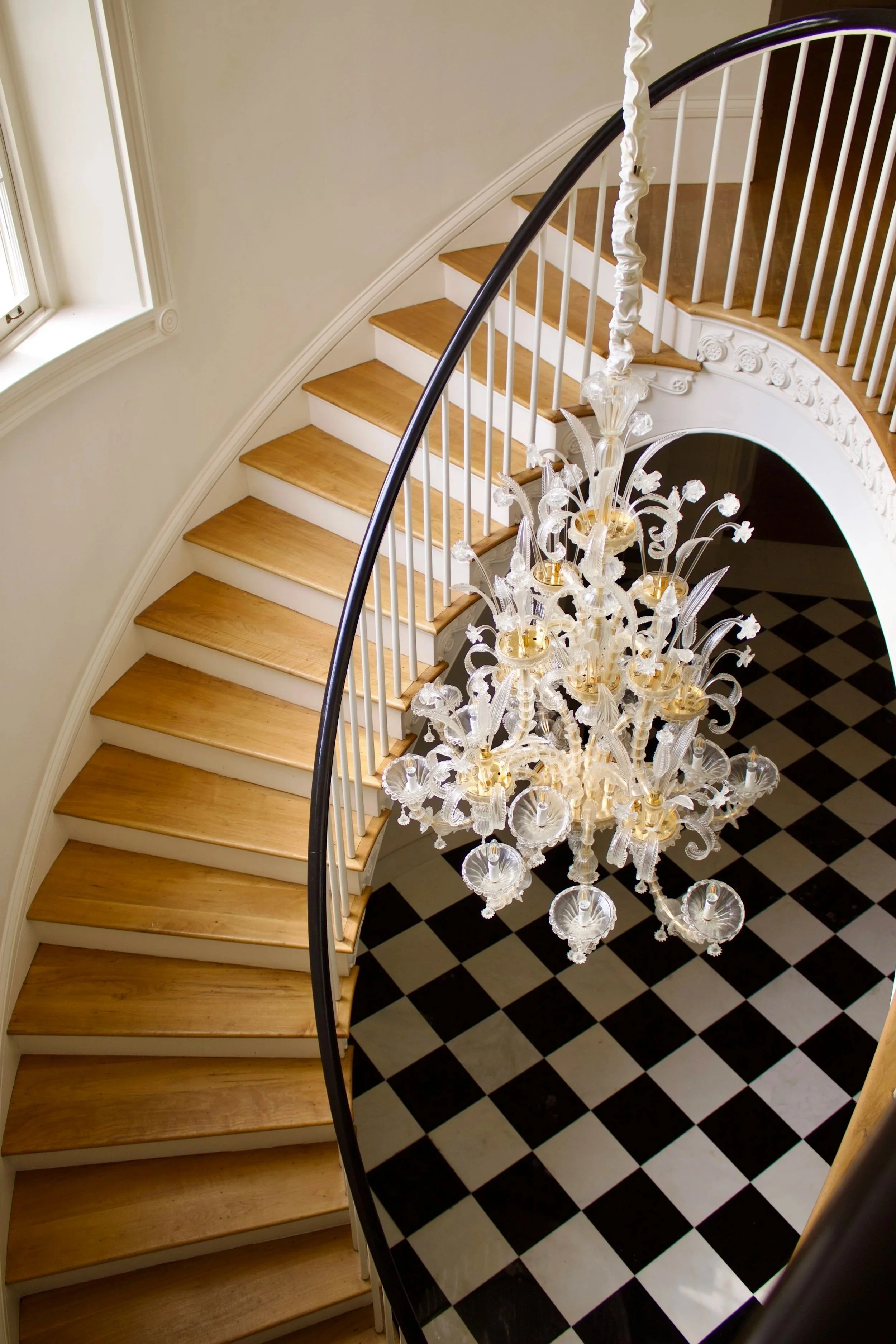 View from top of staircase showing a black and white checkered floor, a curved staircase with wooden steps and white railings, a large ornate crystal chandelier hanging from the ceiling, and a window on the left wall.