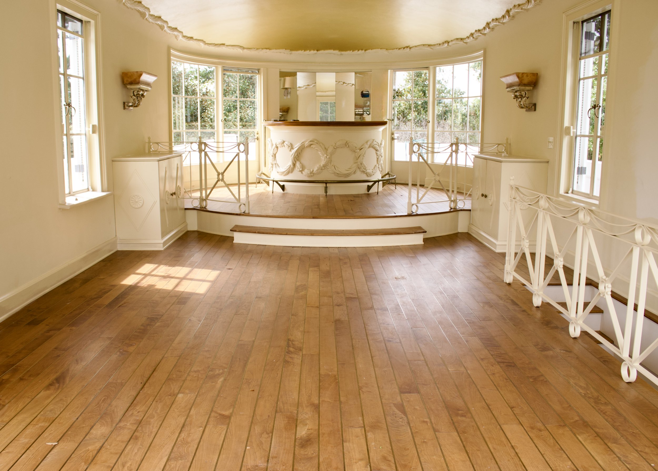 Empty, well-lit room with wooden flooring, large windows, and a curved railing. There is a small stage area in the center with decorative railings and a mirror behind it.
