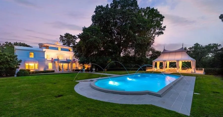 Luxury house with a backyard featuring a swimming pool with fountains, a gazebo, and a large tree, during twilight.
