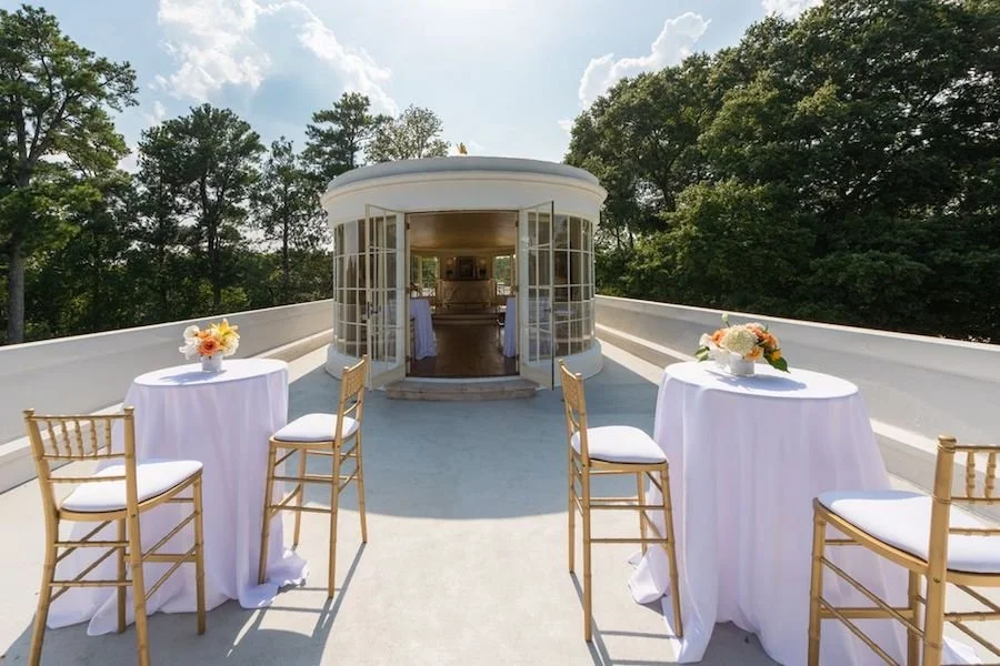 Outdoor rooftop setting with two round tables covered with white tablecloths, each with a floral centerpiece, and four gold chairs with white cushions, overlooking trees and a partly cloudy sky.