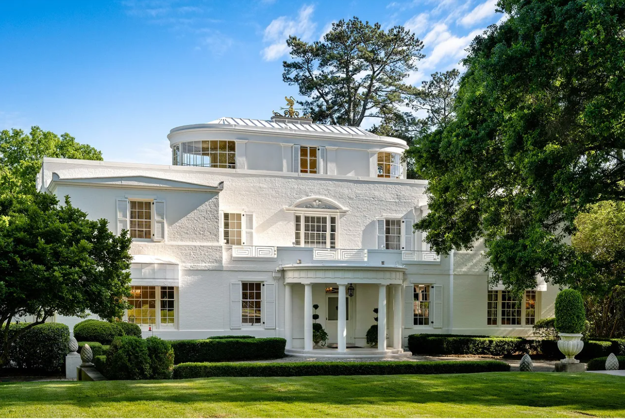 A large, white, multi-story house with an elaborate entrance, surrounded by trees, neatly trimmed bushes, and a well-maintained lawn, under a partly cloudy sky.