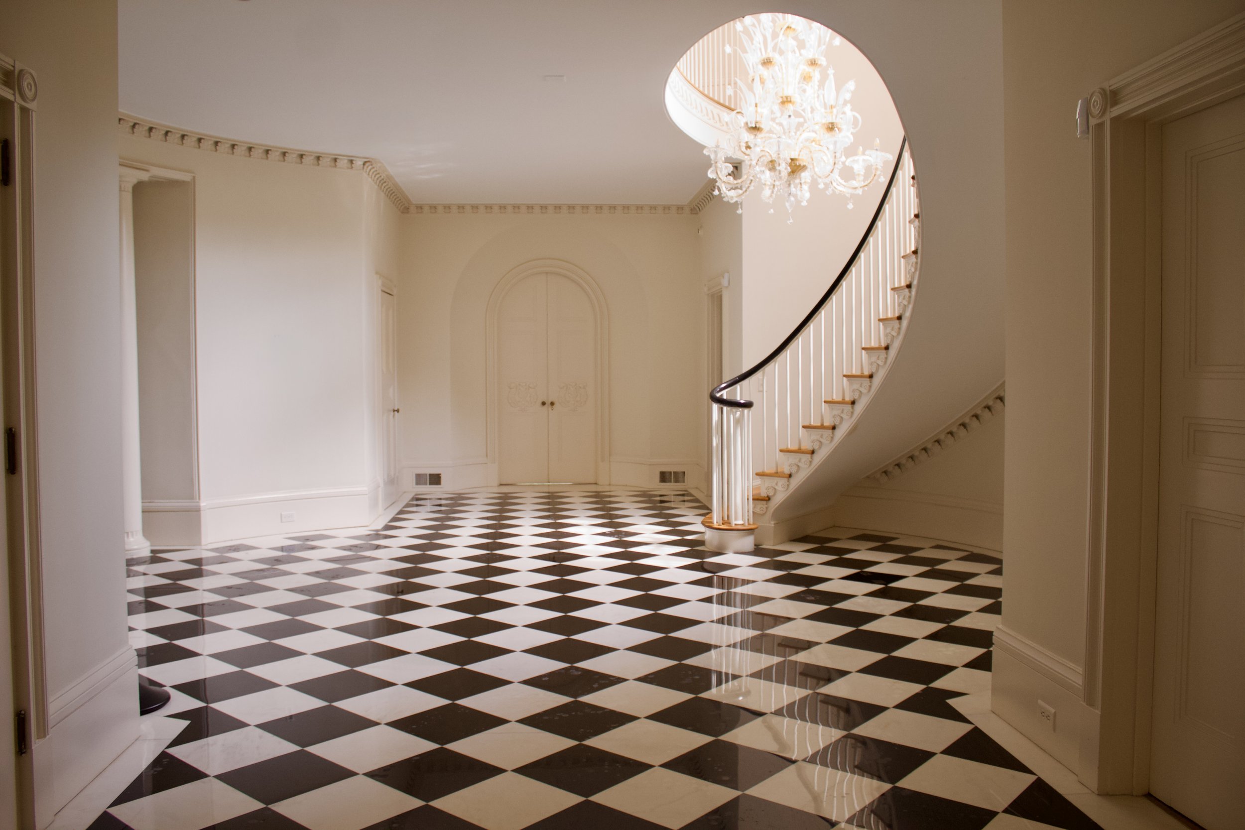 Elegant, spacious foyer with black and white checkered marble floor, white walls, a chandelier, and a curved staircase.