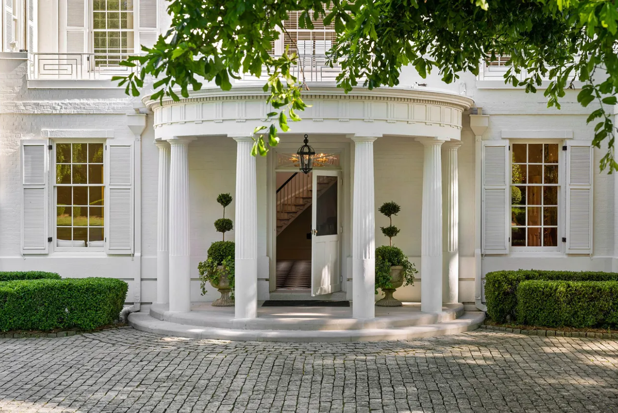Front entrance of a white house with a portico supported by classical columns, surrounded by shrubbery and potted topiary, with a cobblestone pathway leading to the door.