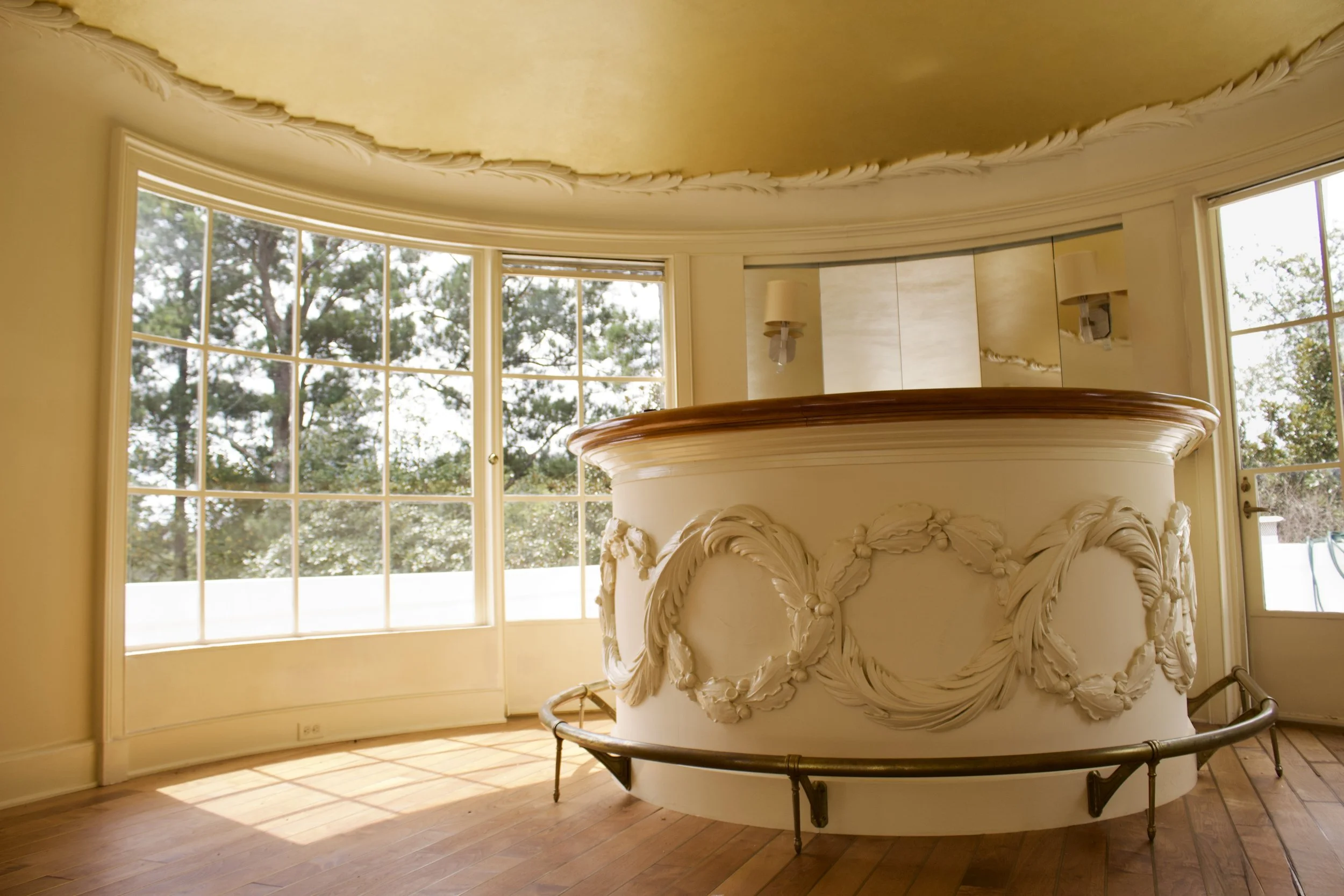 Interior of a room with large curved windows, a decorative ceiling with moldings, and an ornate bar or counter with sculpted garlands. The room is lit by natural light from the windows.