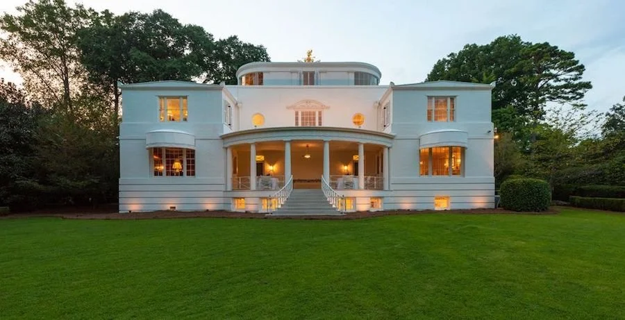 Large white mansion with curved balcony, rounded roof structure, steps leading to front door, surrounded by a green lawn and trees, with warm lighting inside.